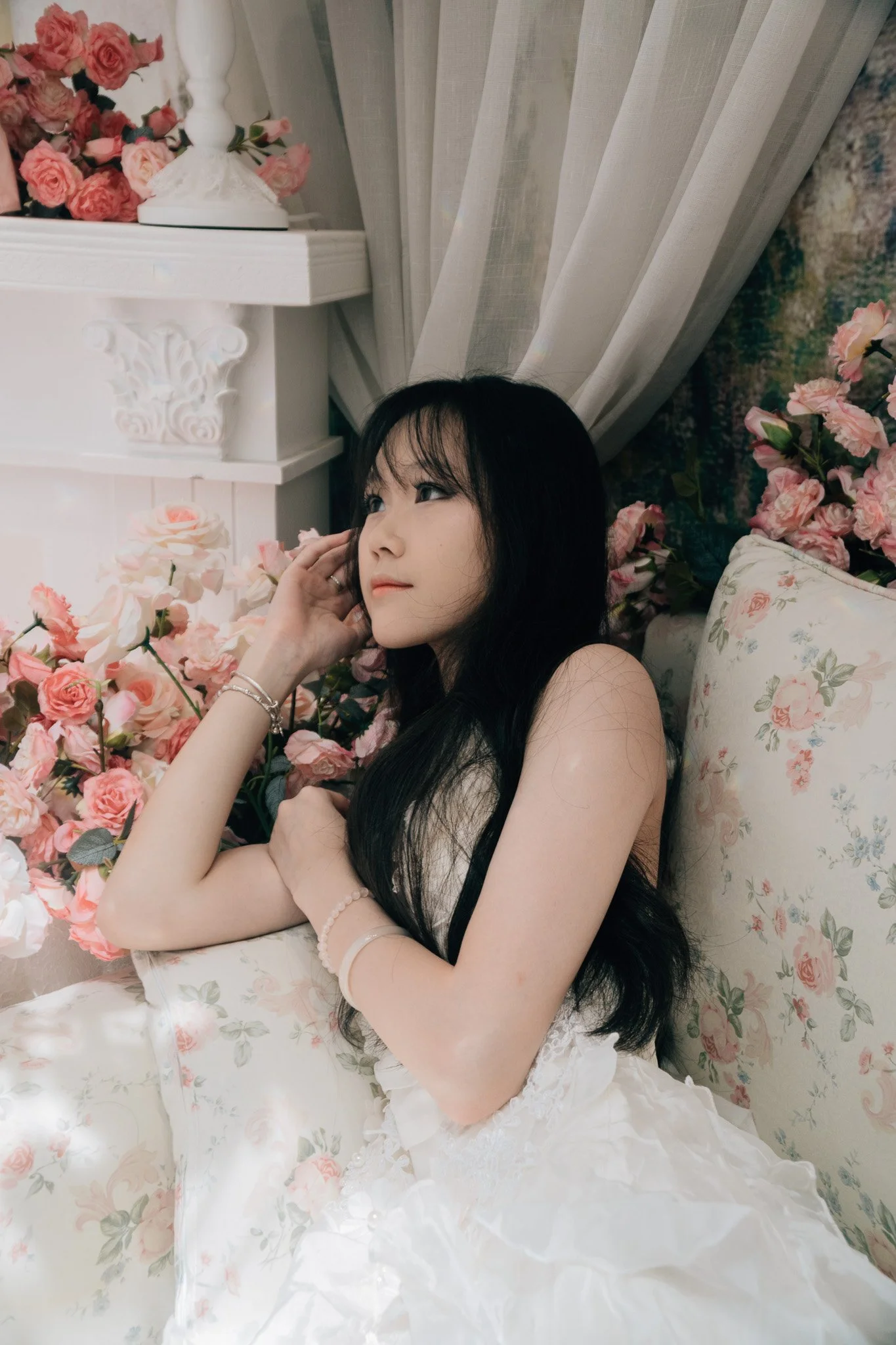 A young woman with long black hair lies on a floral-patterned bed surrounded by pink roses. She is wearing a white dress with lace details and has jewelry on her wrist and fingers. The room has white curtains and a white shelf with a lamp and more pi