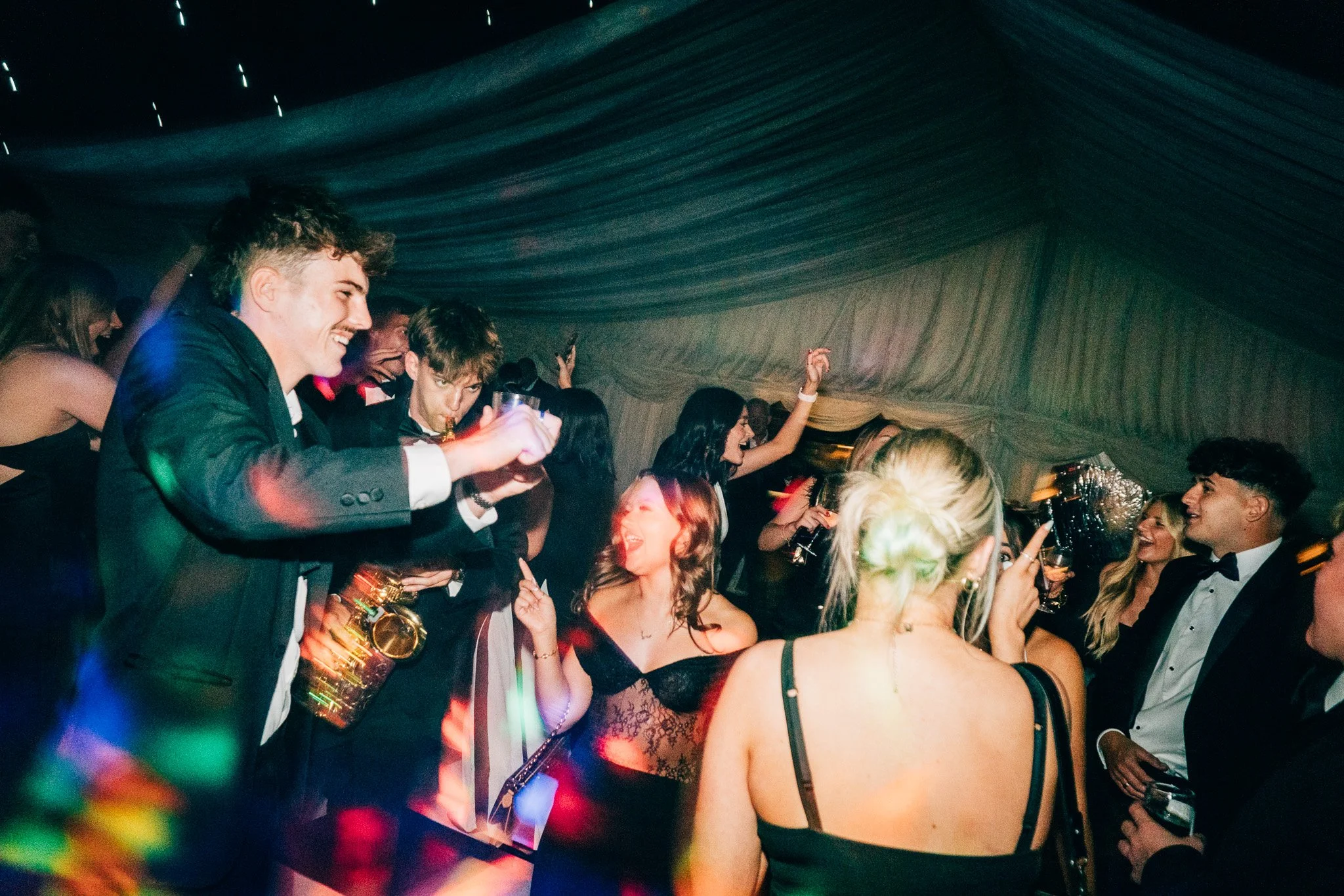 People dancing and celebrating at an elegant party, with some holding drinks and enjoying music under a decorated ceiling.