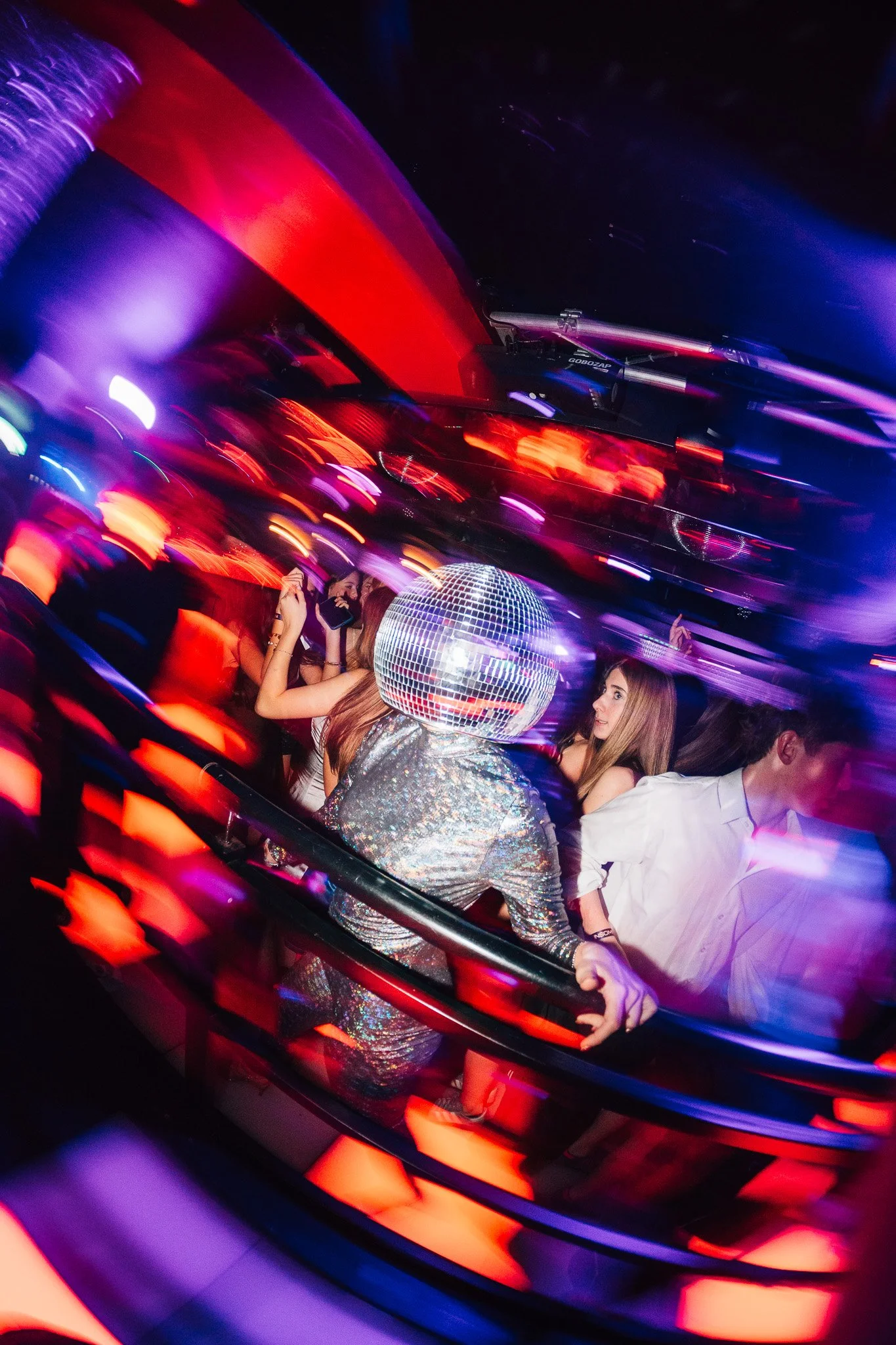 People dancing at a nightclub illuminated by colorful lights, with a disco ball overhead and motion blur effects.