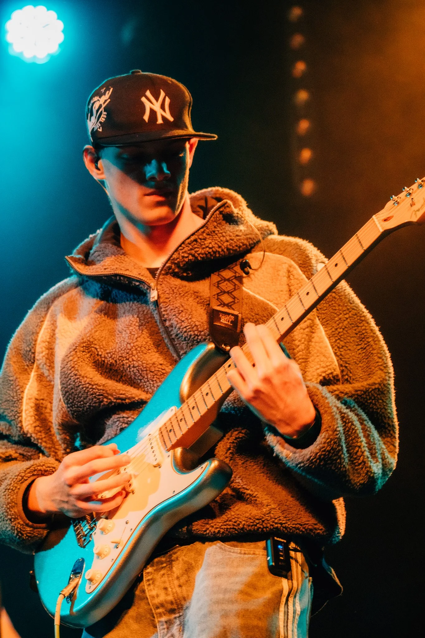 A young man wearing a New York Yankees cap and a fleece jacket is playing an electric guitar on stage, illuminated by blue and orange stage lights.