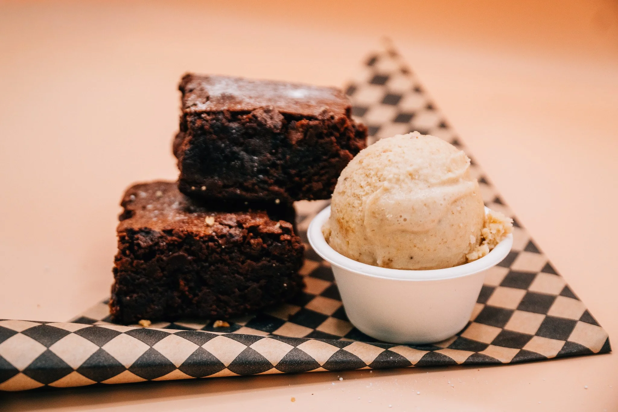 Three chocolate brownies and a scoop of vanilla ice cream served in a small white cup, on checkered food tray.