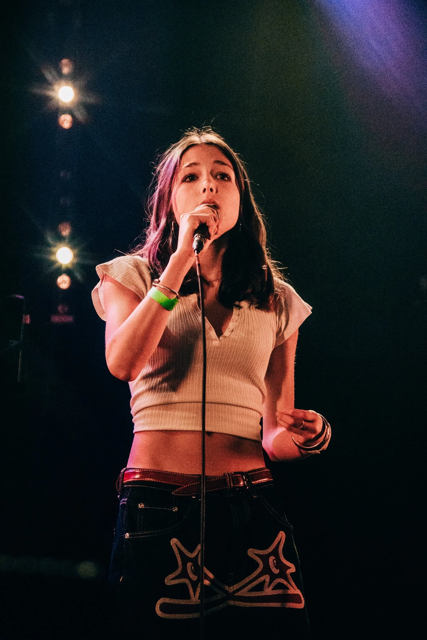 A young woman with long dark hair singing into a microphone on stage, wearing a beige cropped top and black pants with star designs, illuminated by stage lights against a dark background.