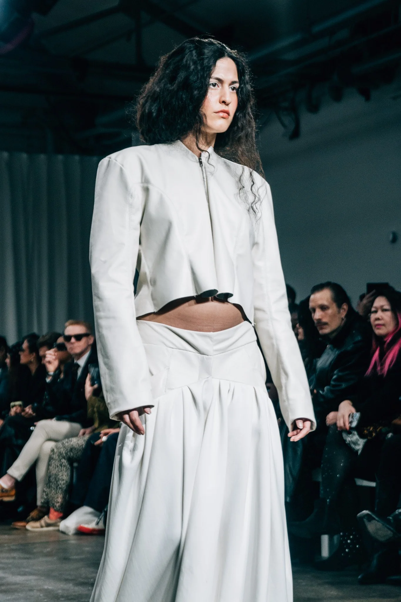 A female model with curly black hair walking on an indoor runway, wearing a white cropped jacket and white wide-leg pants, with an audience seated on either side of the runway.