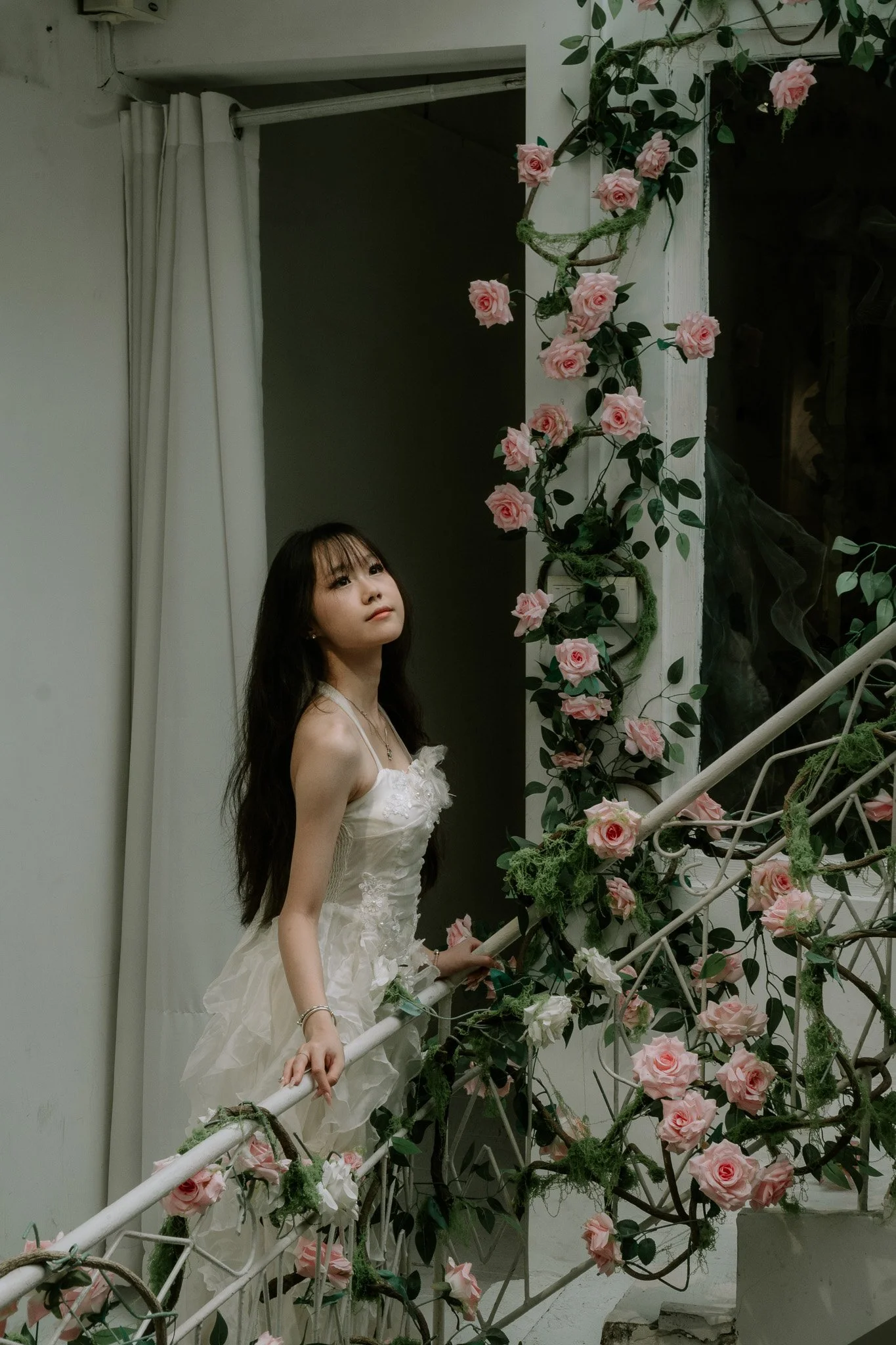 A young woman in a white dress standing beside a decorated railing with pink roses and greenery, indoors near a white wall and curtain.