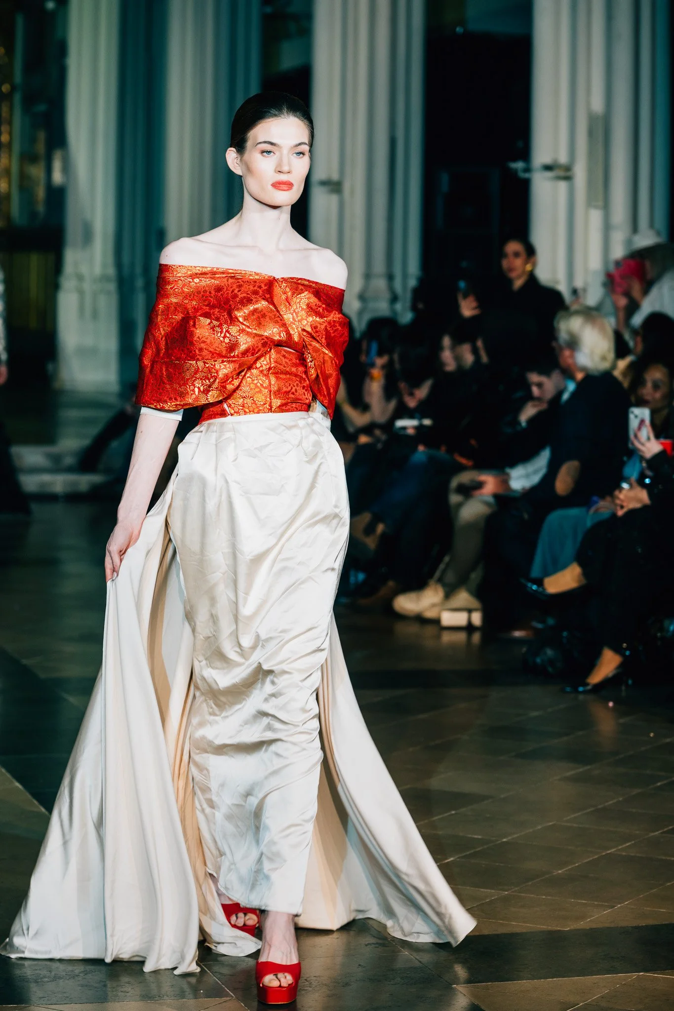 Model walking on runway in a fashion show, wearing a red off-the-shoulder top with gold embroidery, white high-waisted pants, red heels, and bright red lipstick, with audience in background.