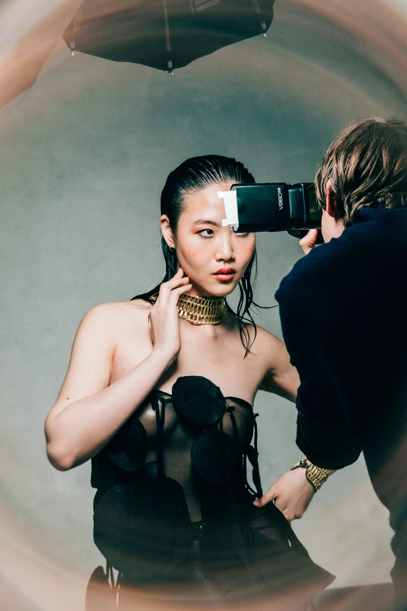 A fashion photoshoot with a female model with wet hair, wearing a black strapless top with pom-pom details, gold jewelry including a chunky necklace and bracelet, and a black sheer fabric with pom-poms. The model is being styled or directed by a pers