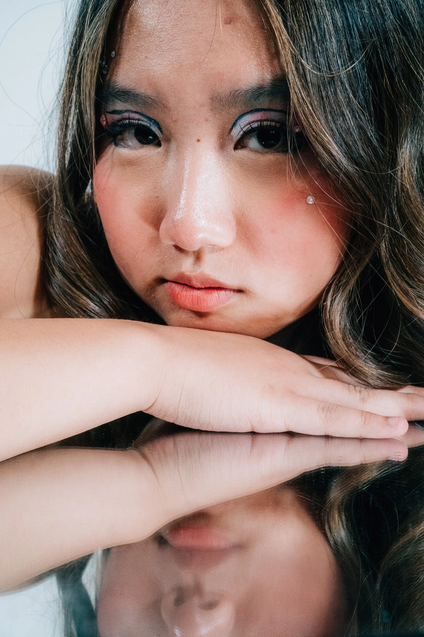 A woman with makeup resting her head on her arm, with long dark hair and a small clear earring.