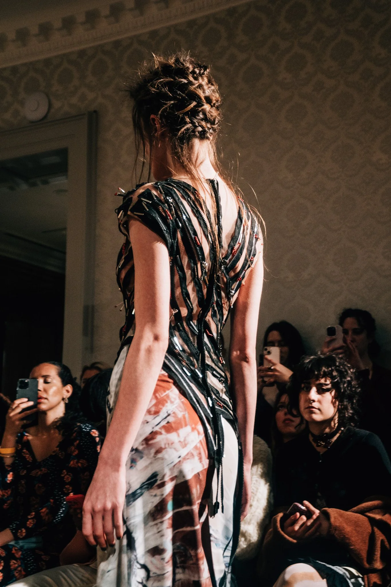 A model walks on a runway wearing a dress with black zipper details and a patterned apron. Spectators are watching and taking photos, with some using smartphones. The picture captures a fashion show event.