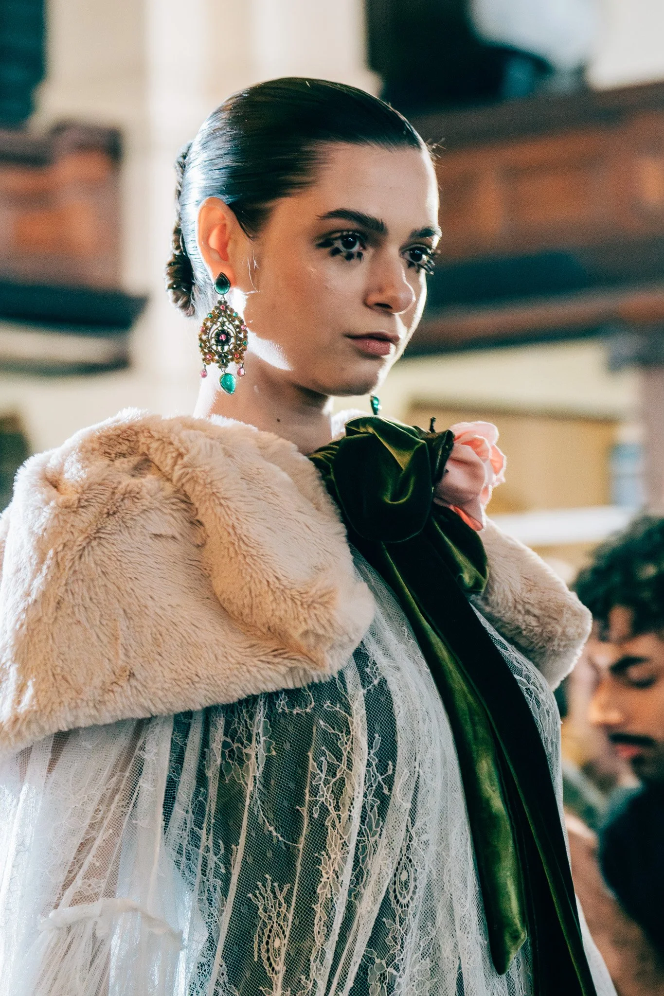 A woman with dark hair styled in a bun, wearing large ornate earrings, a beige faux fur coat over a white lace dress, and a dark green satin ribbon tied around her neck, in an indoor setting.