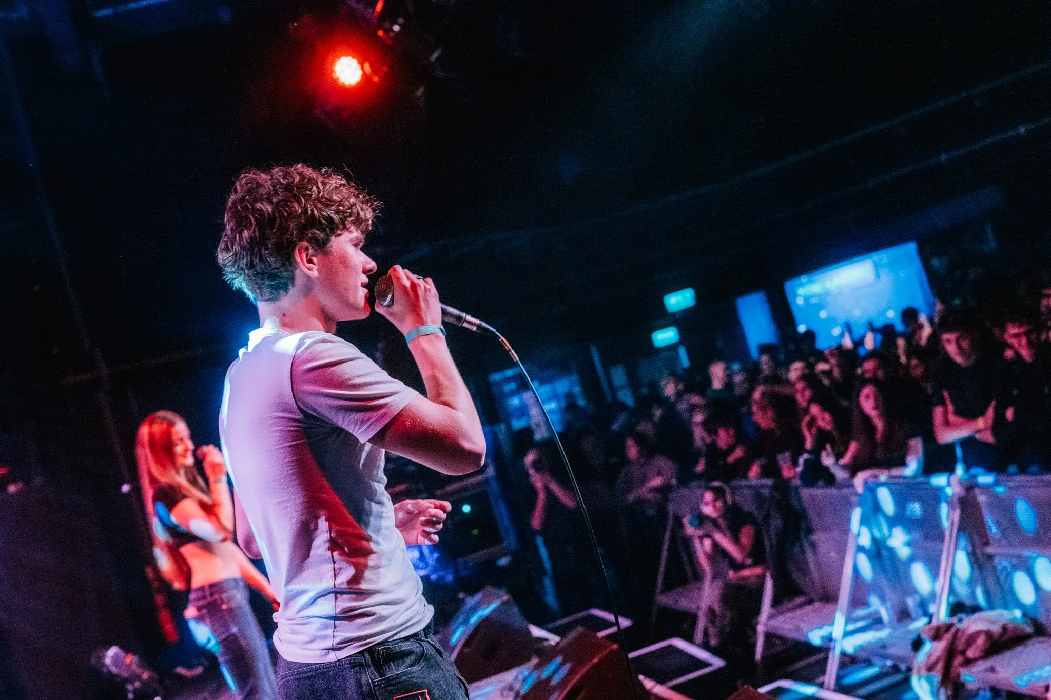 A person with curly hair singing into a microphone during a performance at a nightclub or concert venue, with an audience in the background and blue lighting.