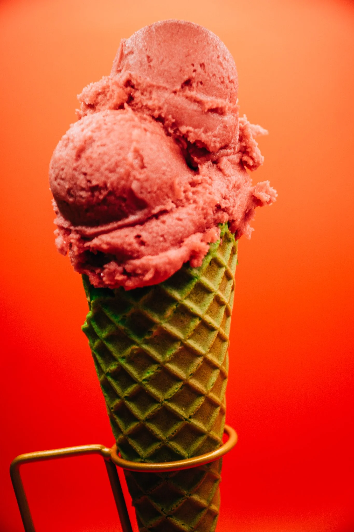 A pink scoop of ice cream in a waffle cone held in a metal holder against a vibrant orange background.