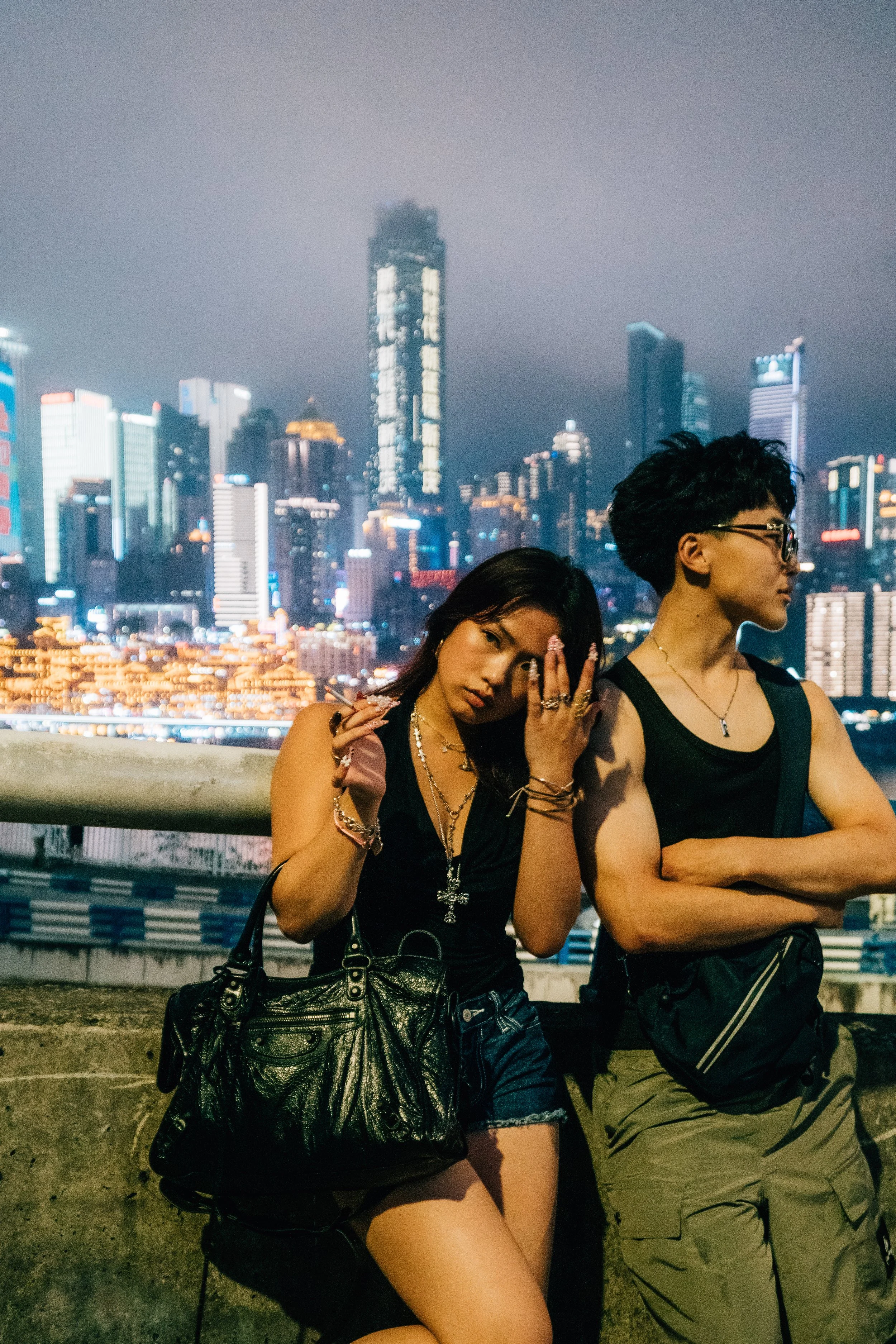 Two young women standing close together in front of a city skyline at night. The woman on the left has long dark hair and is wearing a black sleeveless top, shorts, and accessories including jewelry and a large purse. She is touching her face with on