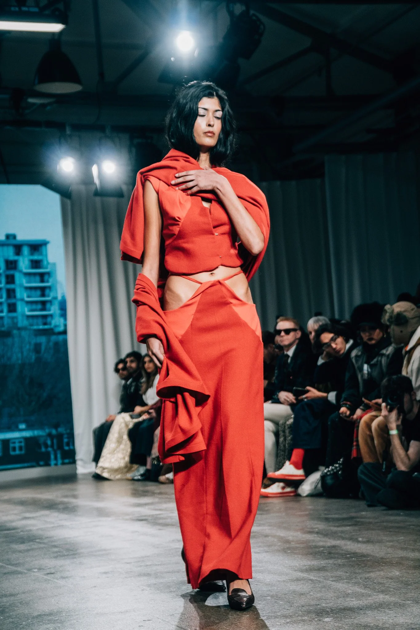 A woman walking on a runway wearing a red outfit with black shoes, with an audience seated on both sides and bright lights overhead.