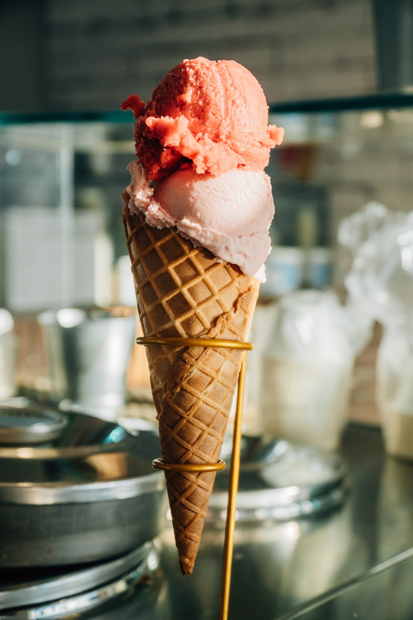Close-up of a double scoop ice cream cone with strawberry and vanilla flavors, held in front of a stainless steel kitchen background.