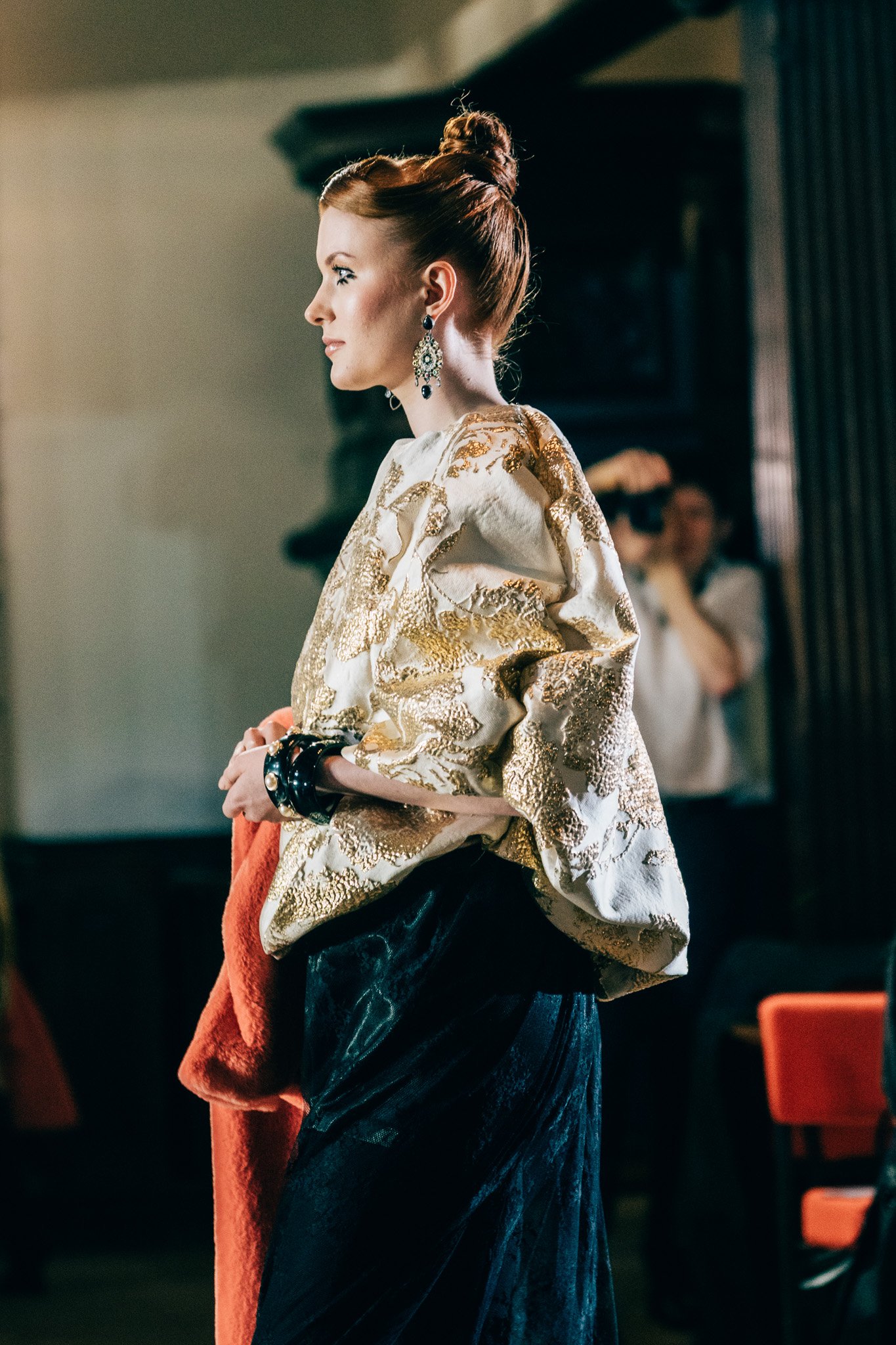 A woman with red hair styled in an updo, wearing ornate earrings and a cream-colored jacket with gold embroidery, standing in a room with dark wood furniture and orange chairs.