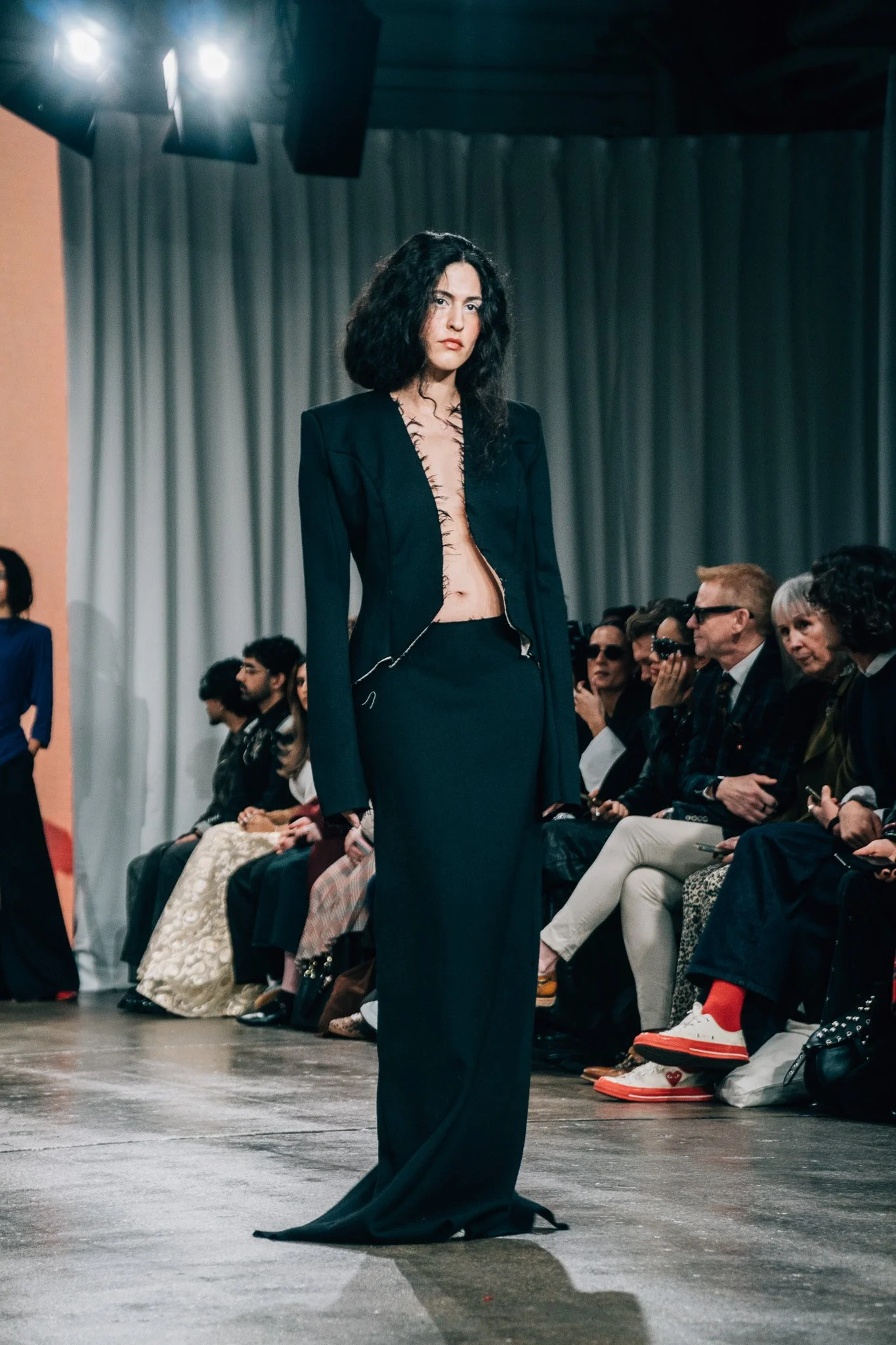 A woman with black curly hair wearing a black blazer and a long black skirt with a deep slit, walking on a runway during a fashion show, with an audience seated on either side.