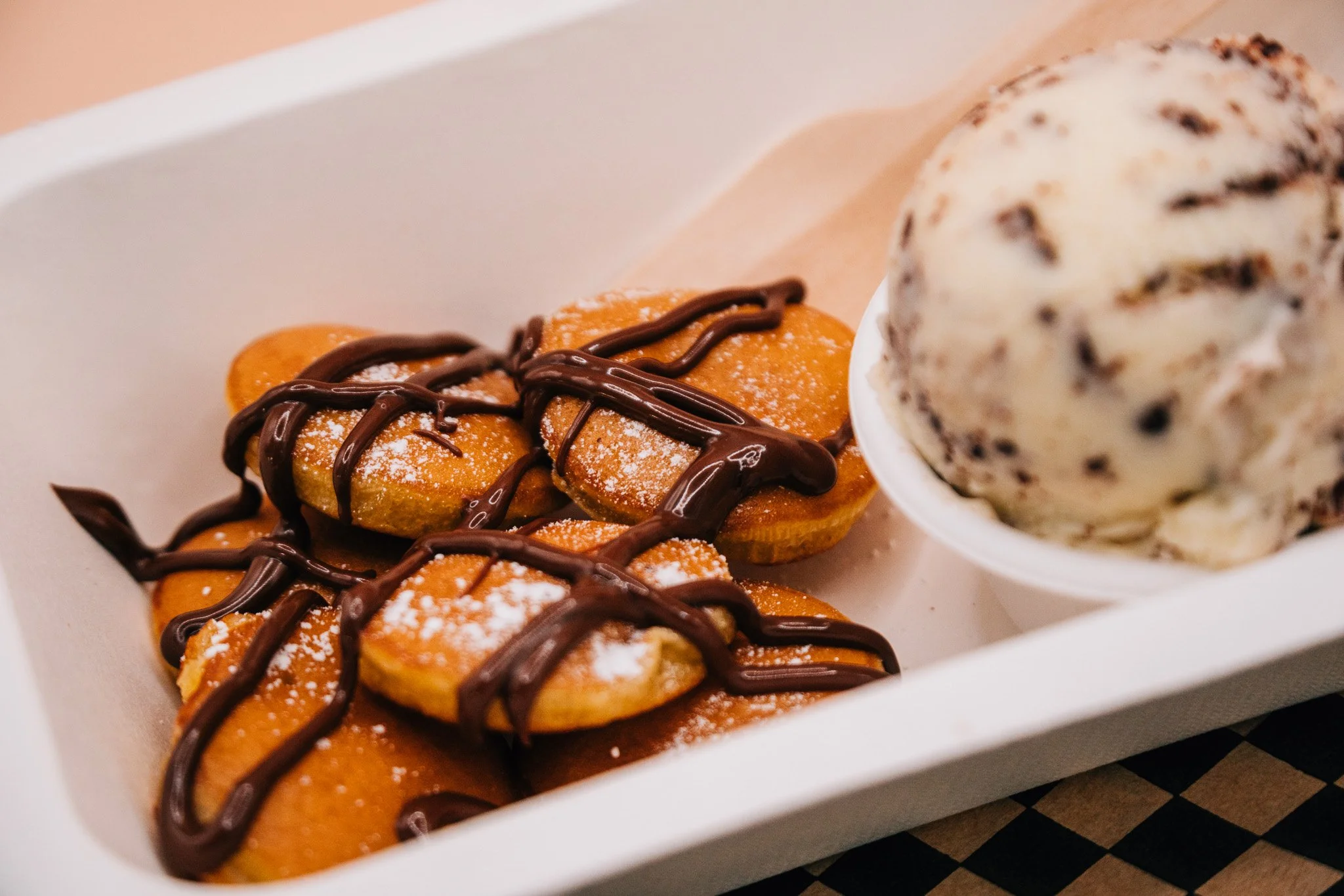A white box contains cookies topped with chocolate drizzle, a small pile of sliced dried apricots, and a scoop of cookies and cream ice cream with chocolate chunks.