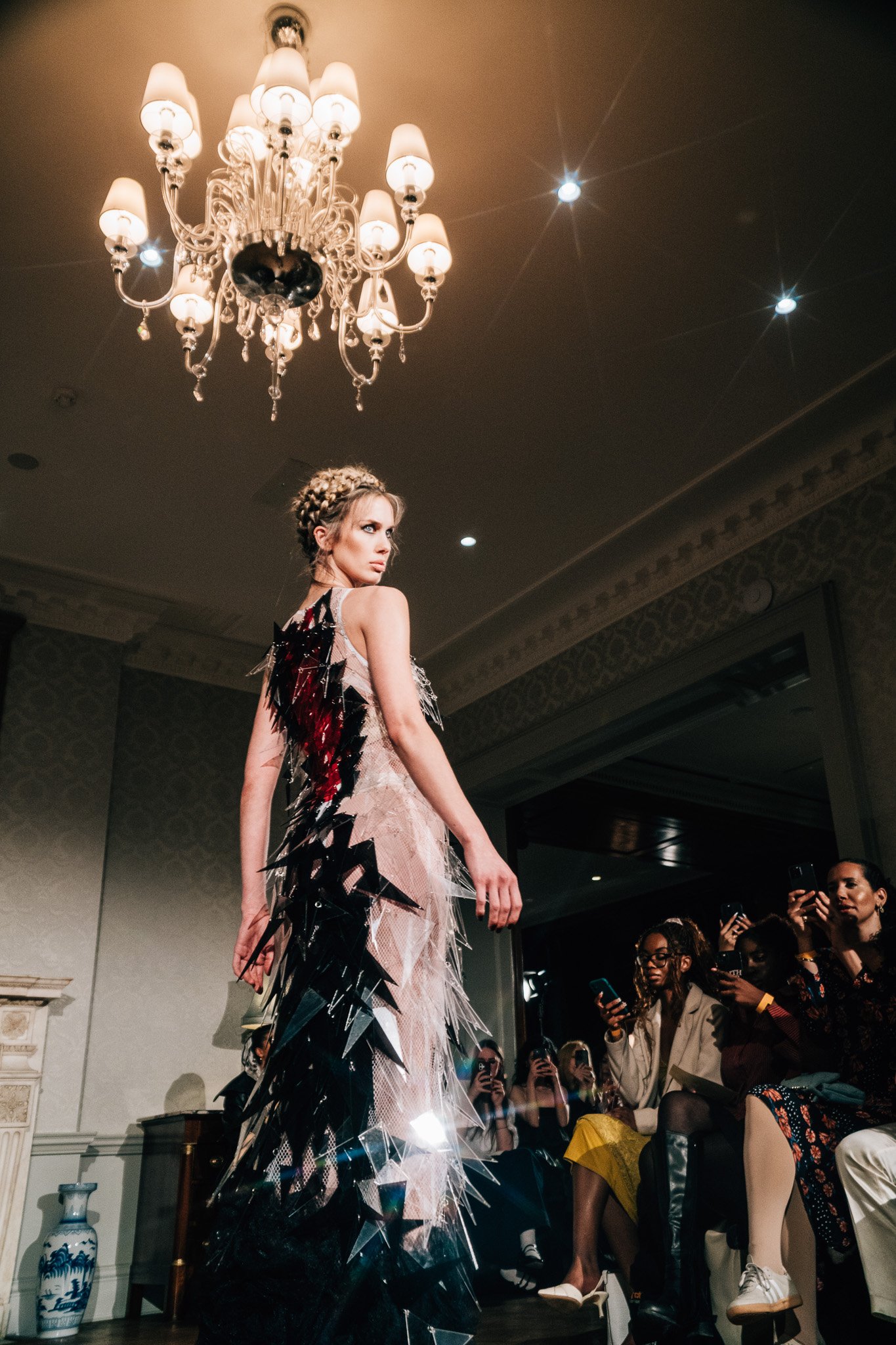 A female model on a runway wearing a dress with black and red feather-like details, during a fashion show with an audience of observers taking photos.