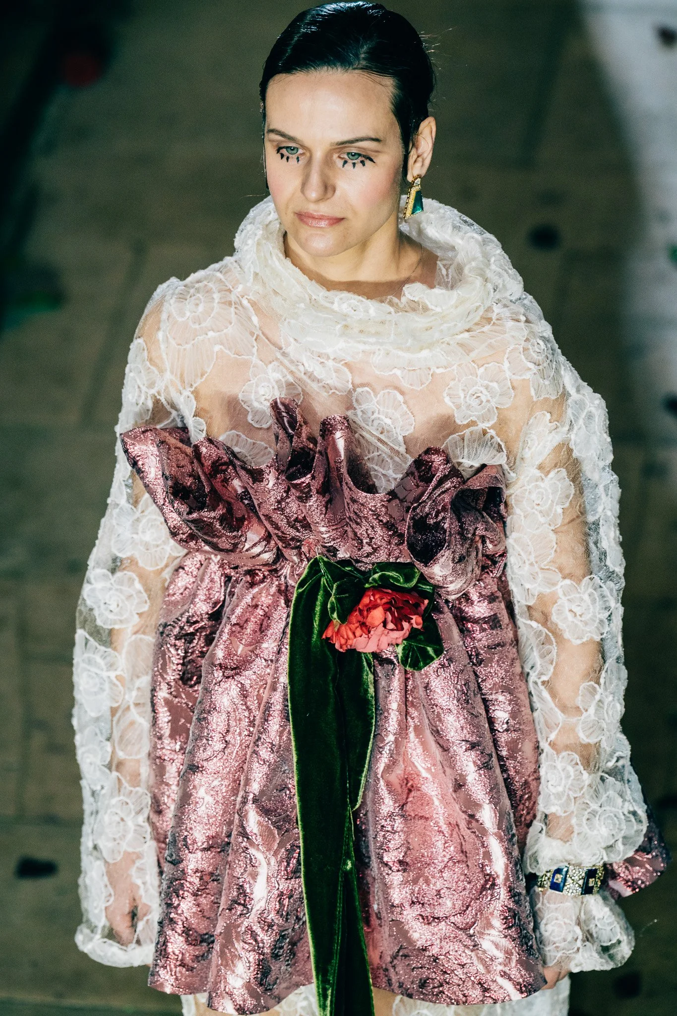 A woman with dark hair styled back, wearing a floral lace dress with a high collar, holding a bouquet of red and pink flowers with green leaves. She has eye makeup with small black dots below her eyes, gold earrings, and a bracelet, and is looking sl