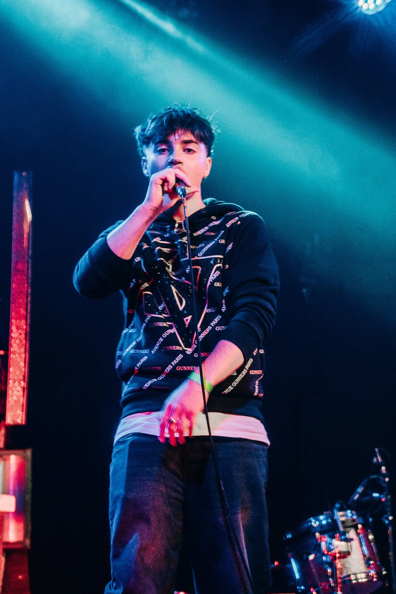 A young man with dark, messy hair performs on a stage, holding a microphone close to his mouth, lit by blue stage lights, wearing a black hoodie with patterned text, white shirt underneath, and dark pants, with musical instruments visible in the back