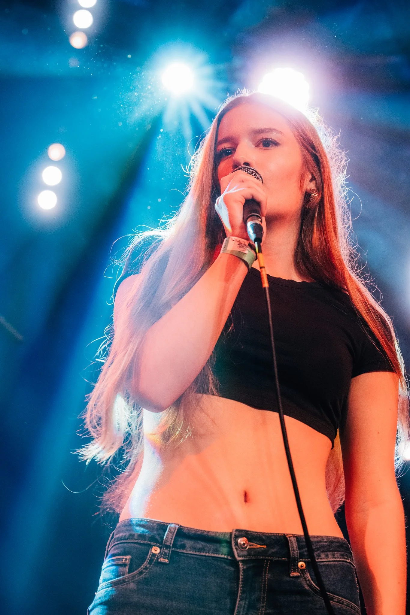 Young woman with long, red hair, wearing a black crop top and jeans, holding a microphone on stage with bright stage lights behind her.