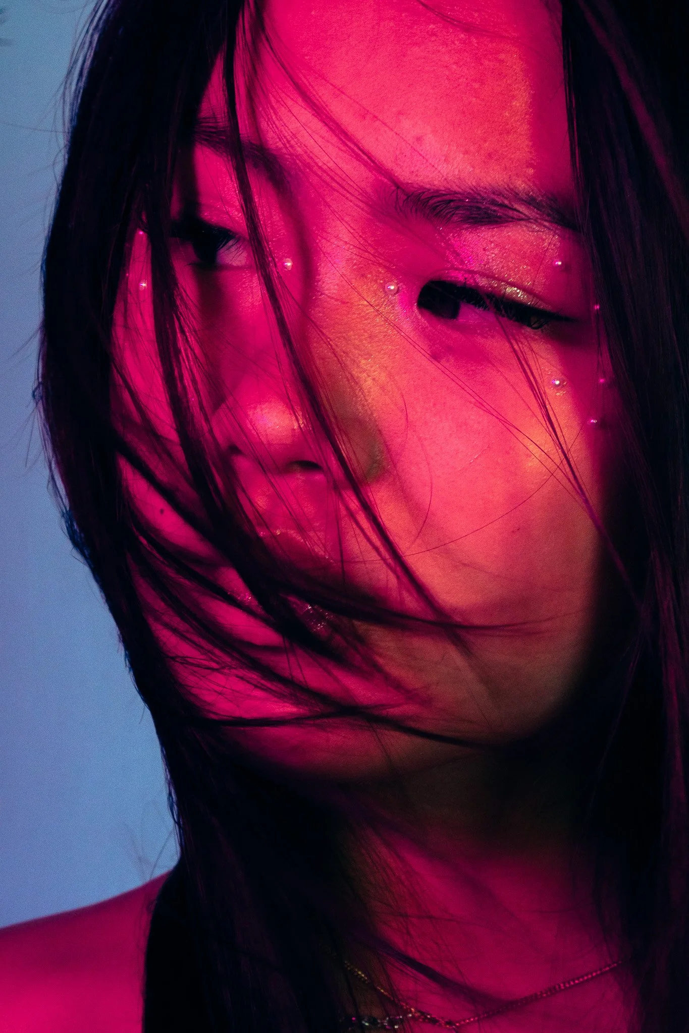 Close-up of a woman's face with black hair, pink lighting, and shiny skin.