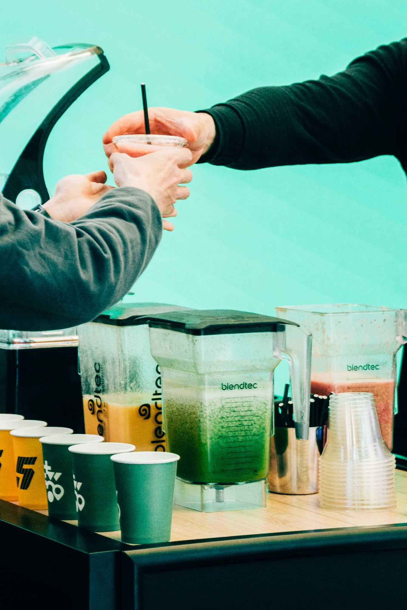 Coffee shop counter with green cups, a blender, and cups of coffee on a wooden surface.