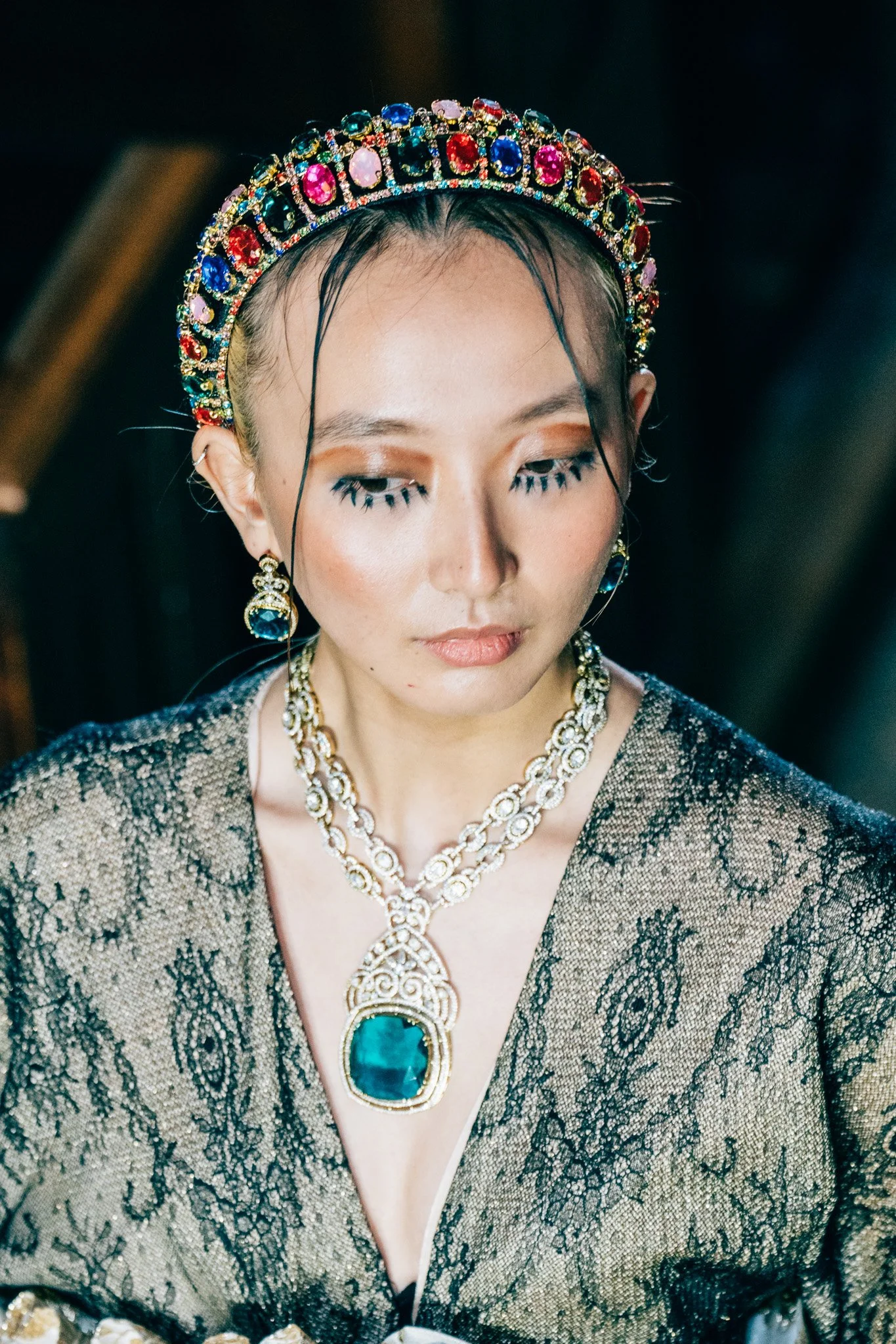 Close-up of a woman wearing a jeweled headband, large gemstone necklace, matching earrings, and an elegant dress with lace detail, with her eyes closed.
