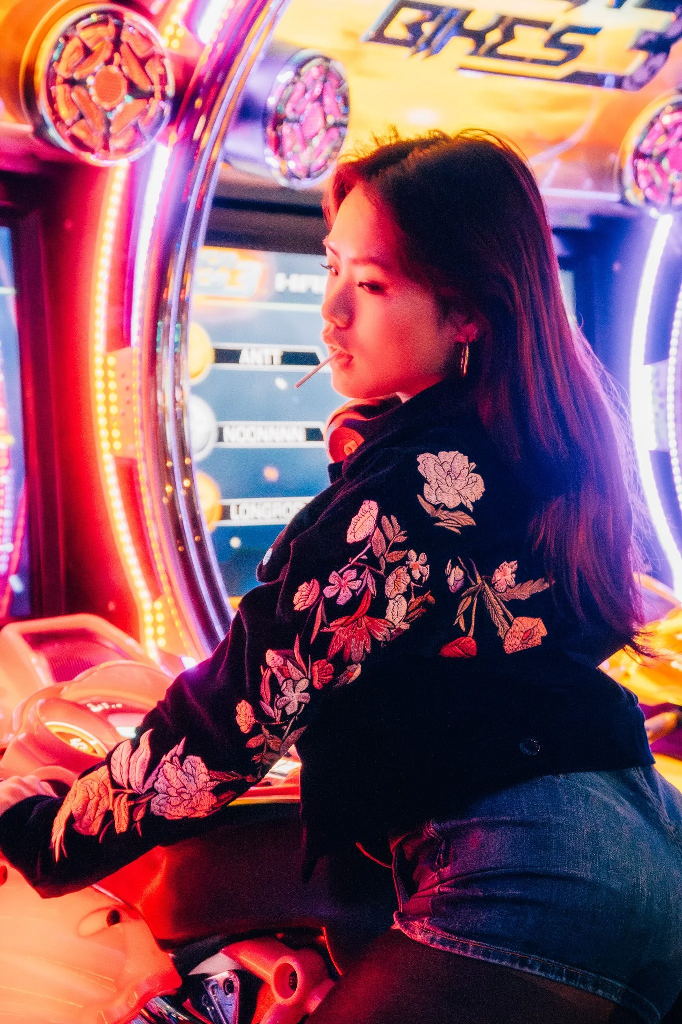 A woman riding an arcade motorcycle game at an amusement center, illuminated by neon lights with a colorful background.