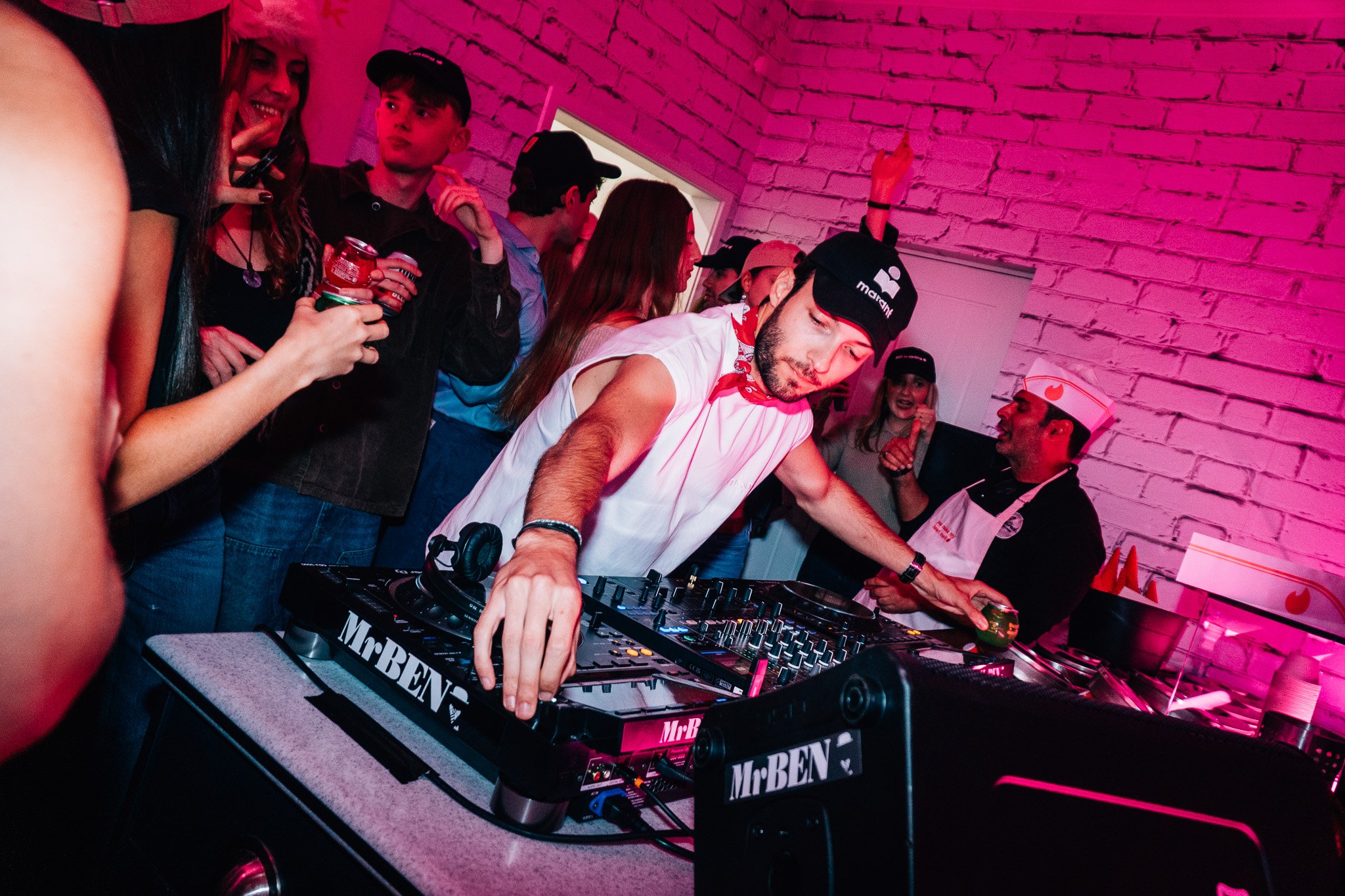 A group of young people at a party, with a DJ mixing music on turntables, in a room with pink lighting and a white brick wall.
