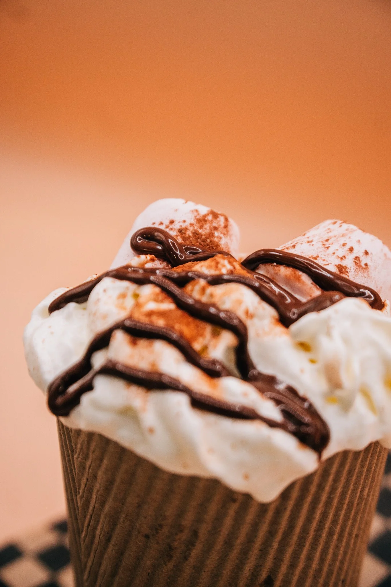 Close-up of a dessert cupcake topped with whipped cream, chocolate drizzle, and sprinkled cocoa powder in a brown paper cup.