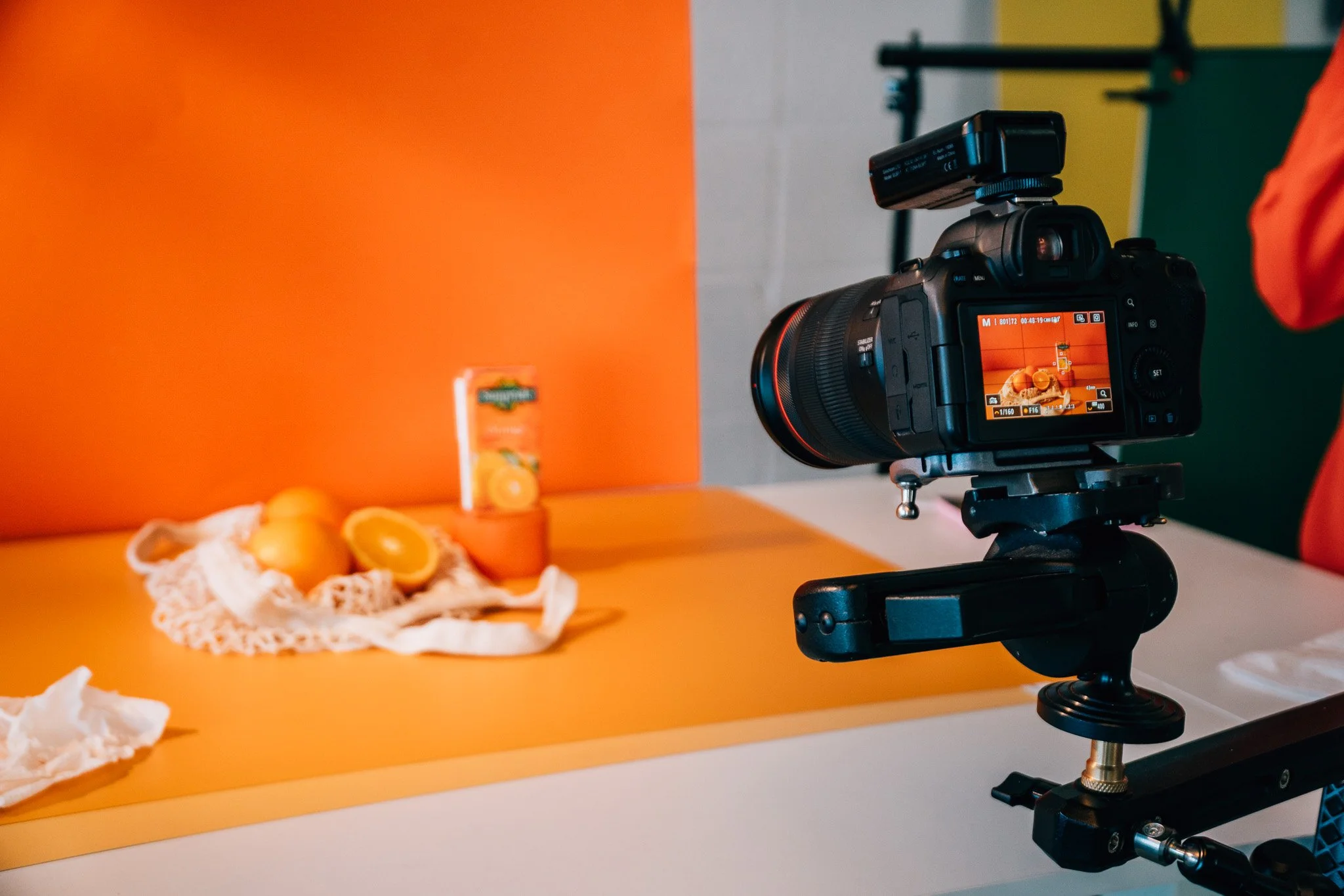 A professional camera on a tripod capturing a photo of oranges and a orange juice carton on a yellow background.