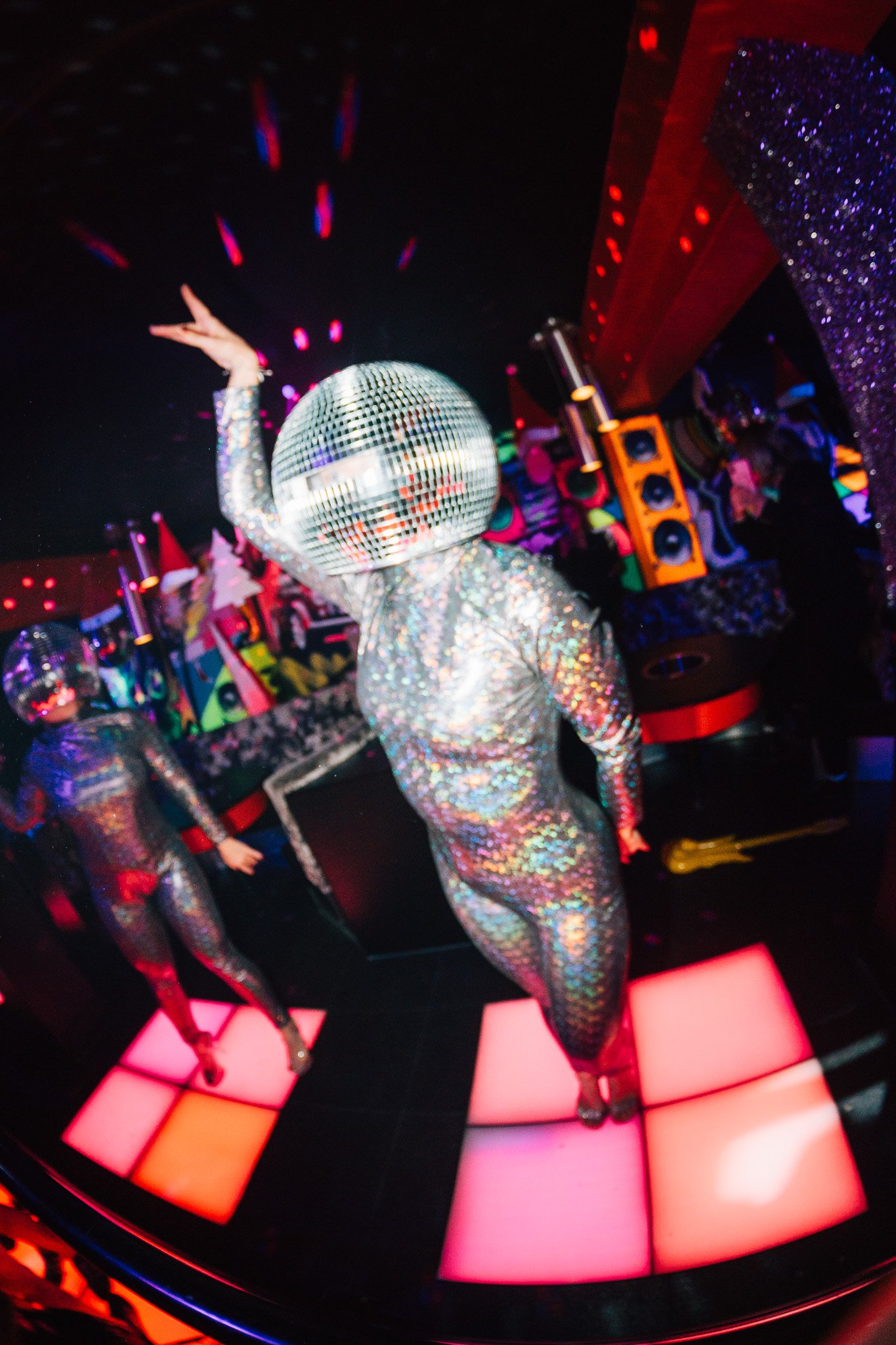 Disco-themed party scene with a person wearing a holographic jumpsuit and a reflective disco ball mask, standing on illuminated pink and orange dance floor tiles with colorful lighting and sound equipment in the background.