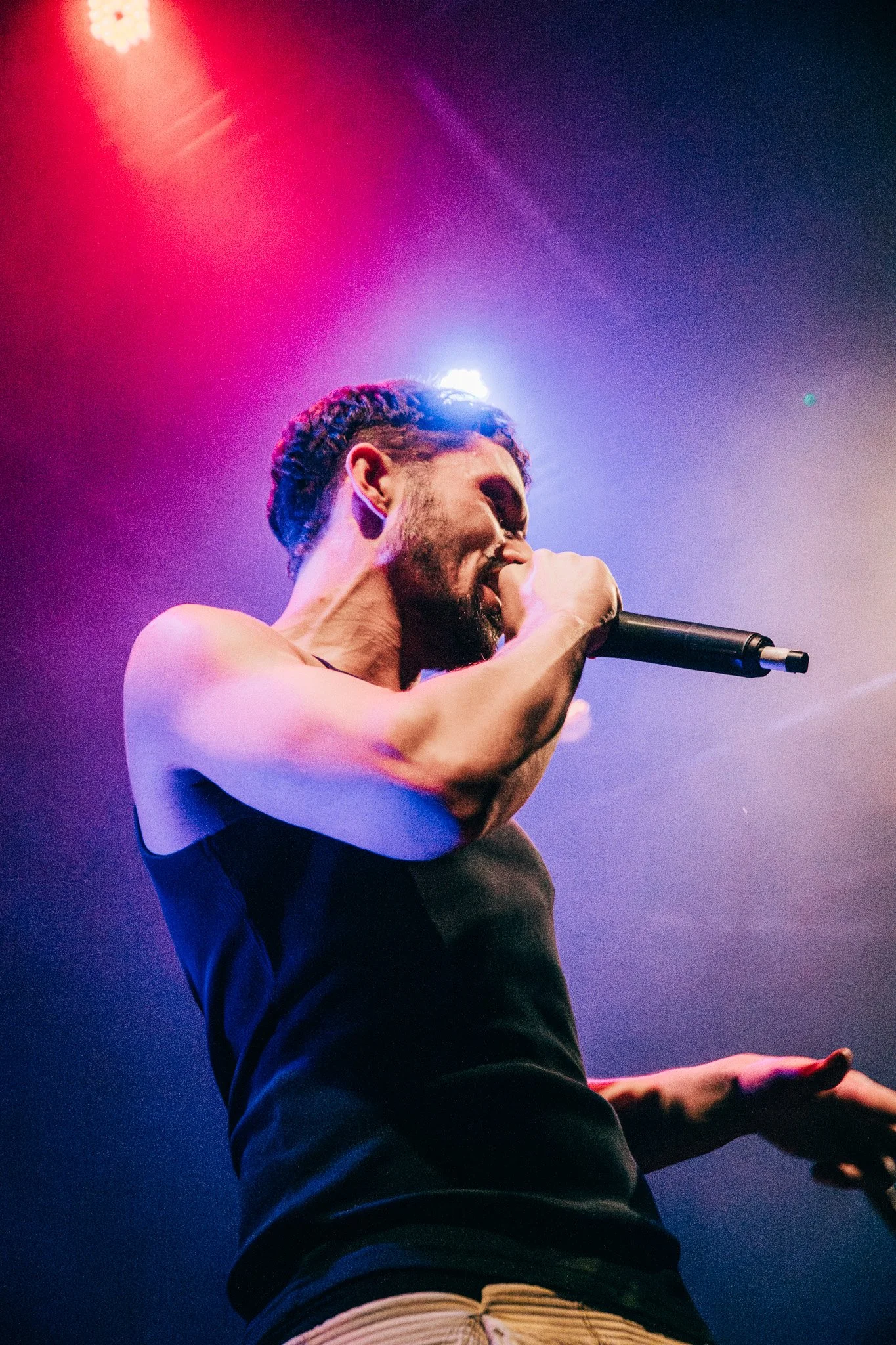 A male performer singing into a microphone under colorful stage lights, with one arm extended and wearing a sleeveless shirt.