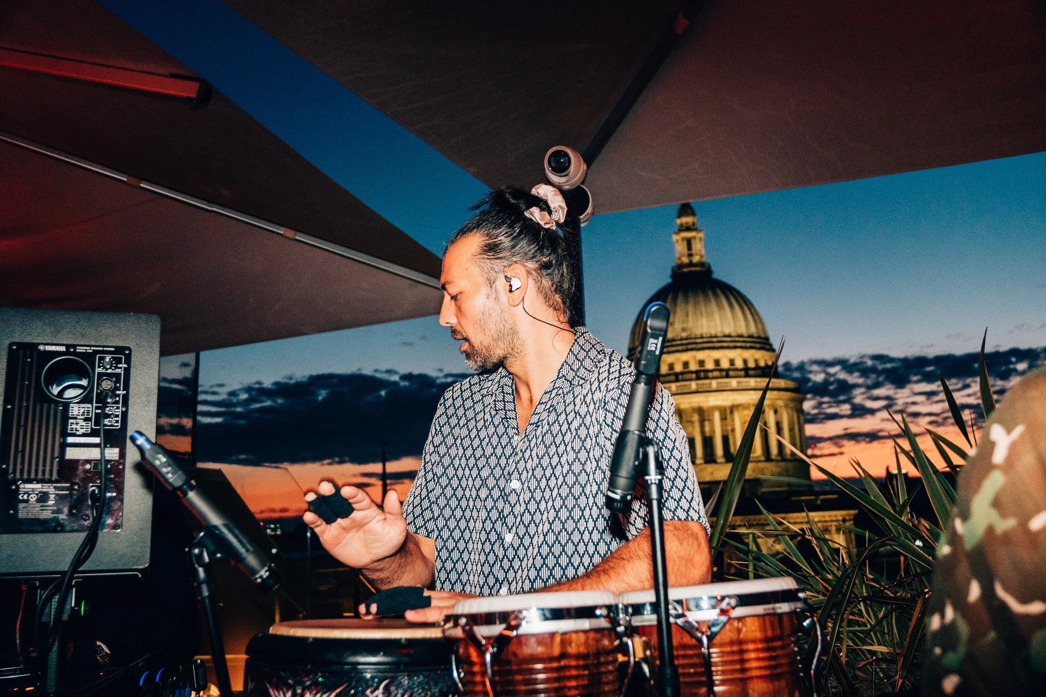 A man with long dark hair tied with a pink scrunchie, wearing patterned shirt, is DJing at an outdoor event during sunset with U.S. Capitol building visible in the background.