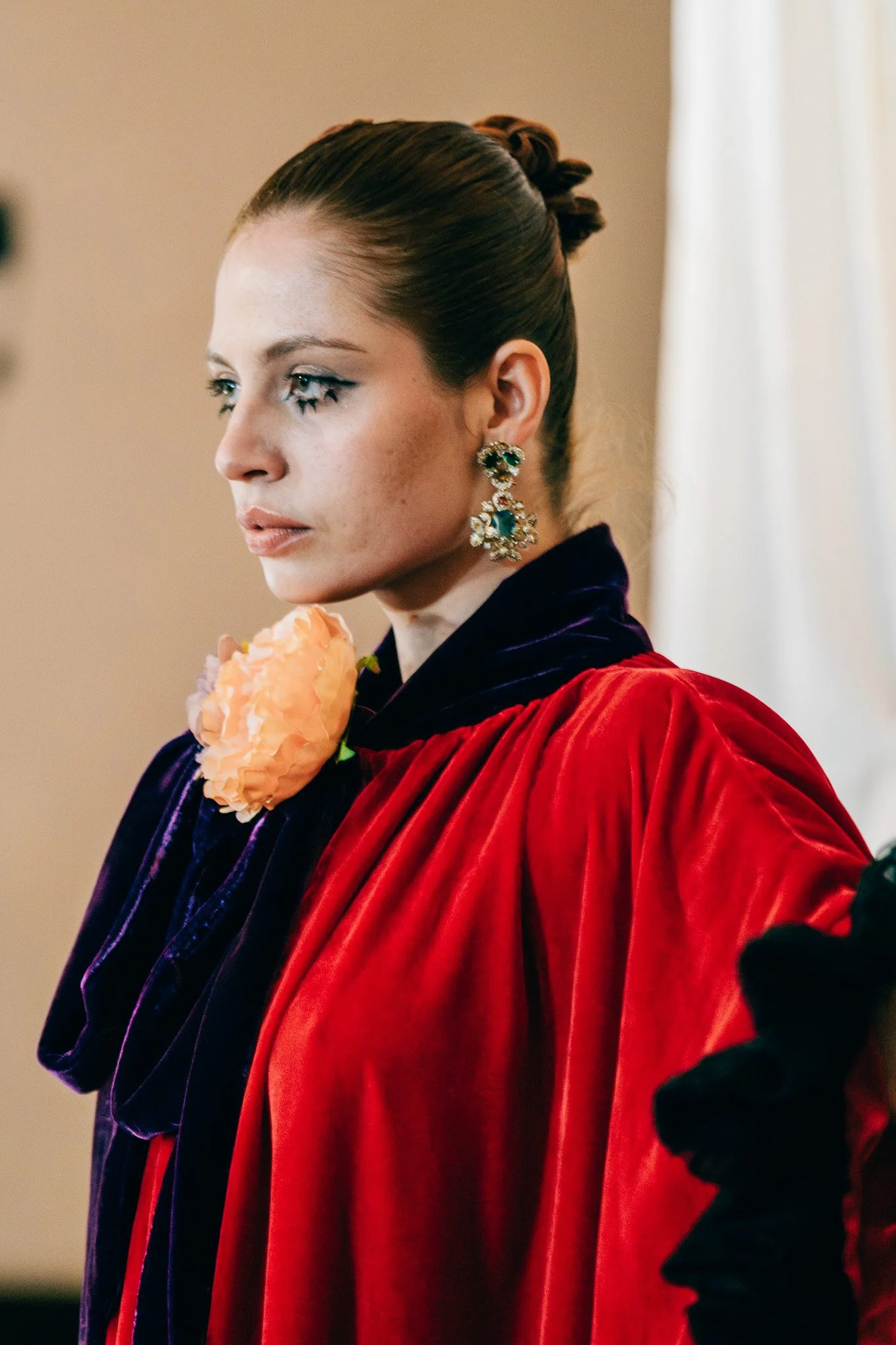 A woman with styled hair and prominent earrings wearing a rich red and dark purple velvet garment, holding a pink flower, standing against a beige background.