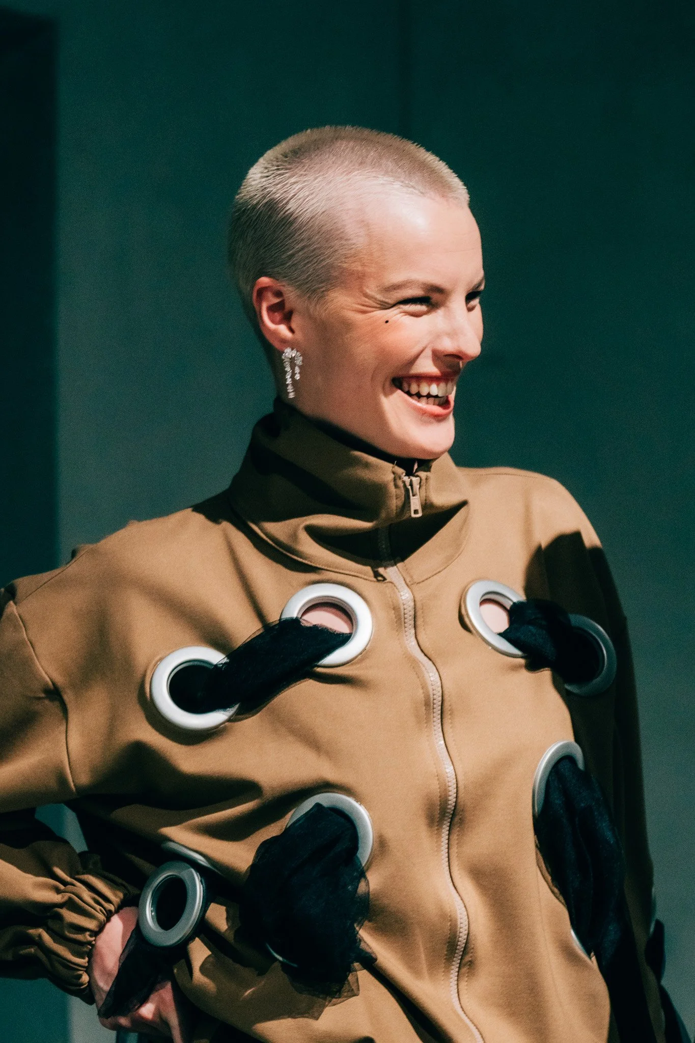 A person with short blonde hair, wearing a tan jacket with circular embellishments on the front, smiling and looking to the right, against a dark teal background.