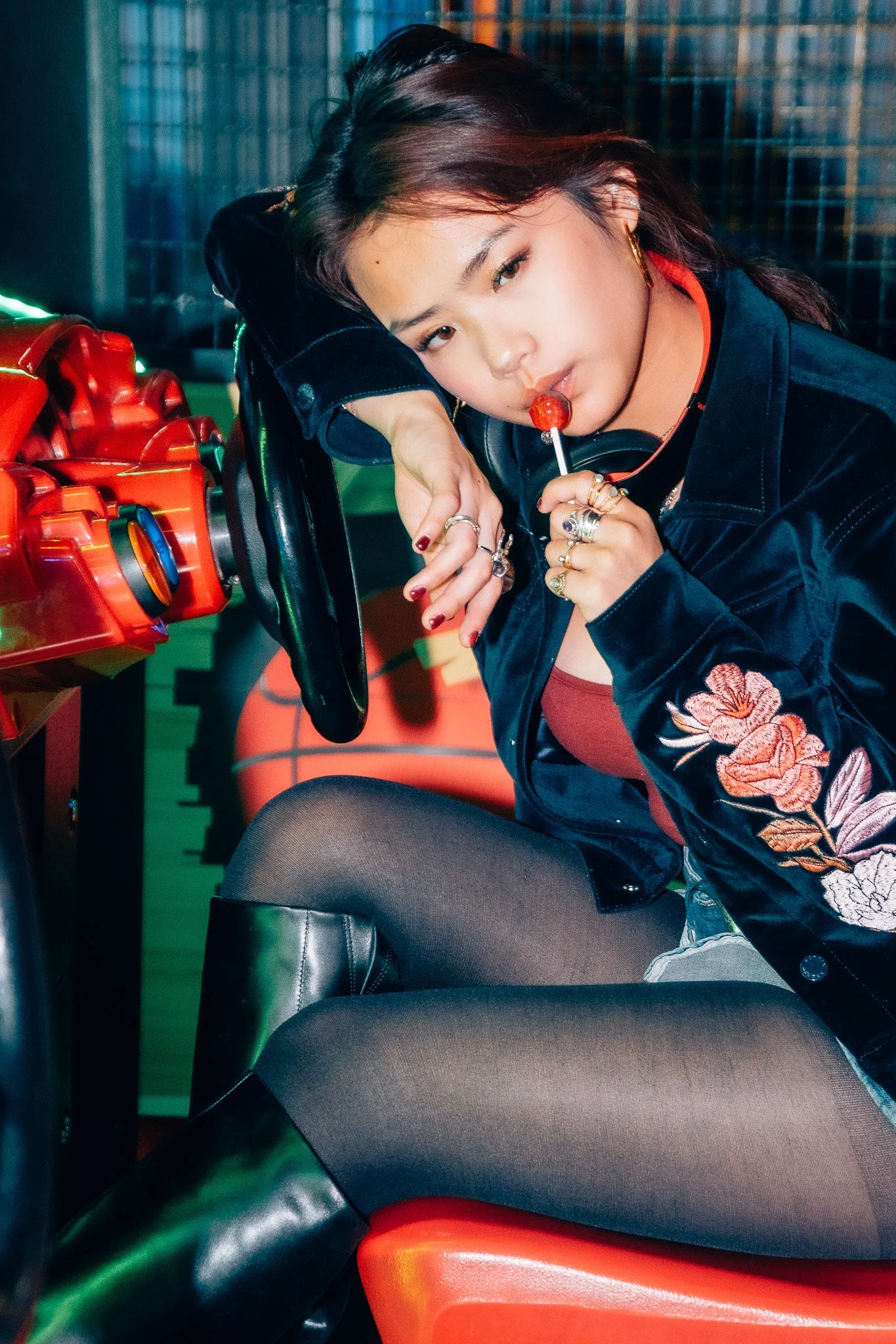 A young woman with dark hair and gold earrings is sitting, resting her head on her arm, holding a red lollipop near her mouth. She is wearing a black jacket with floral embroidery and black leggings, with a green boot visible in the foreground. The b