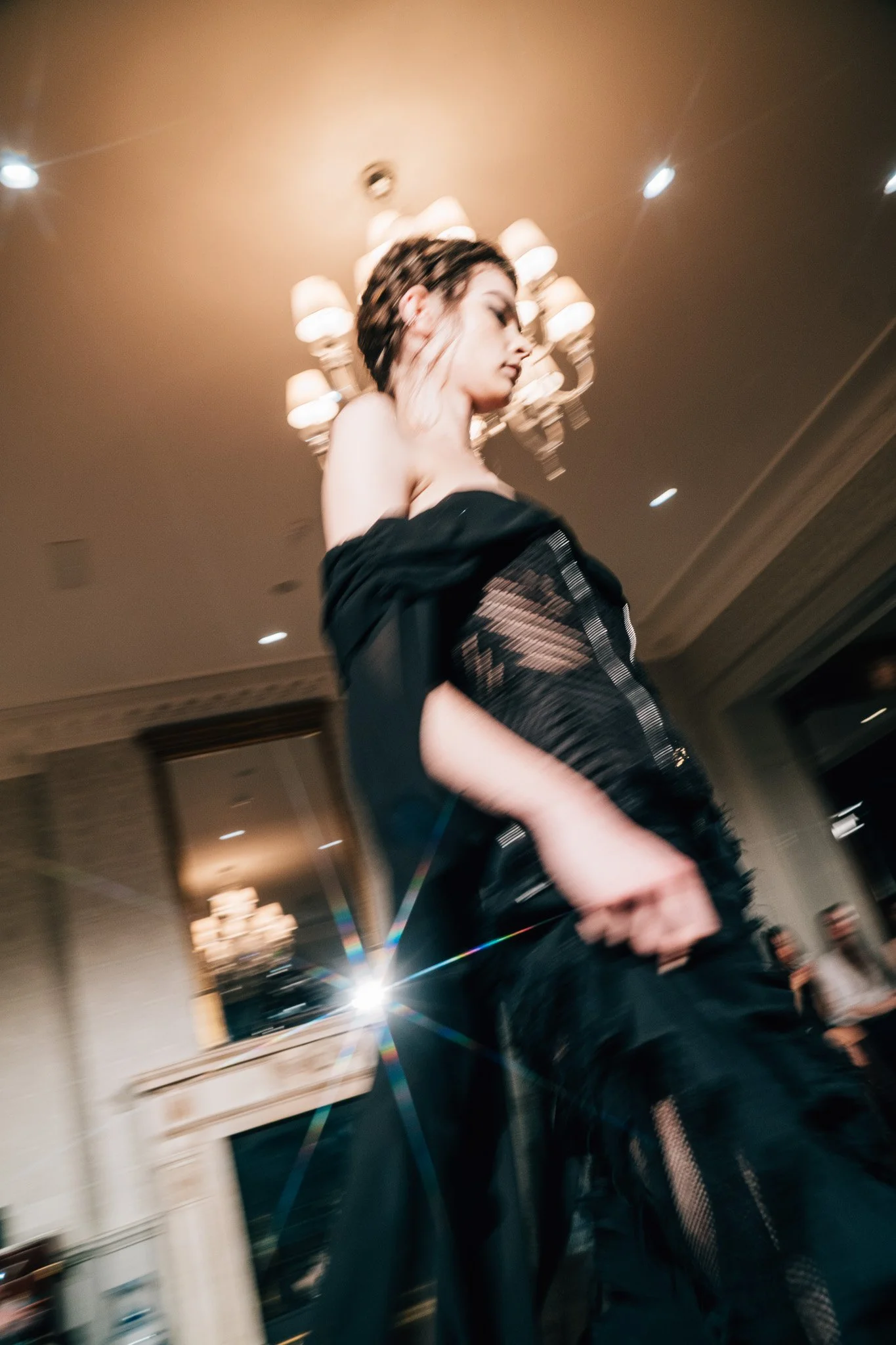 A woman with braided hair wearing a black off-shoulder dress or top, standing in front of a chandelier in an indoor setting.