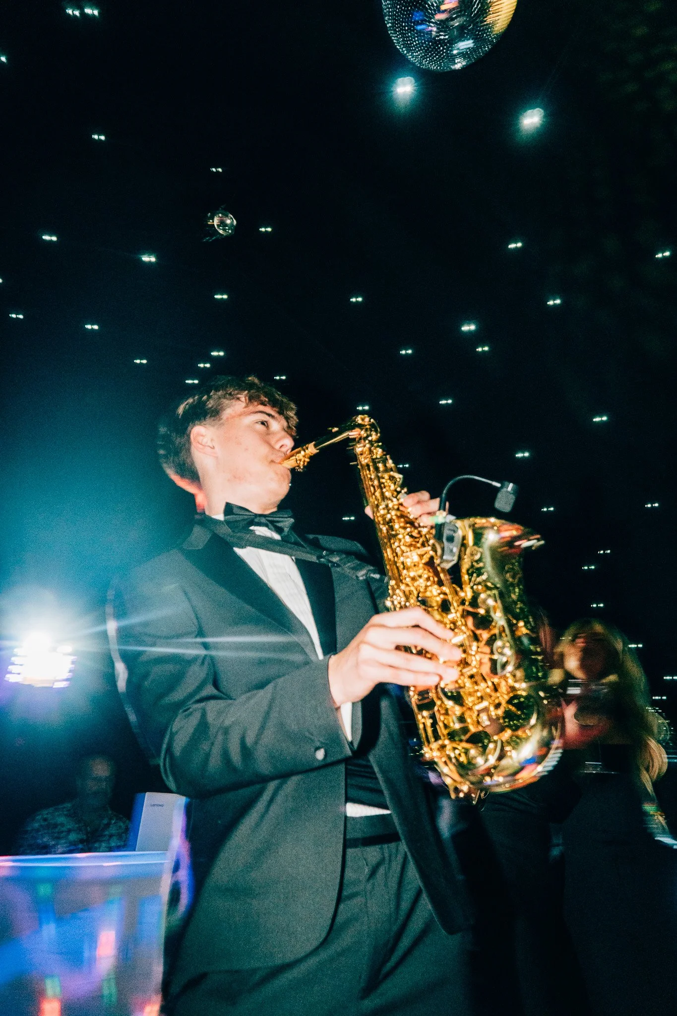 A man in a tuxedo playing a gold saxophone at a dimly lit event with disco balls reflecting lights from the ceiling.