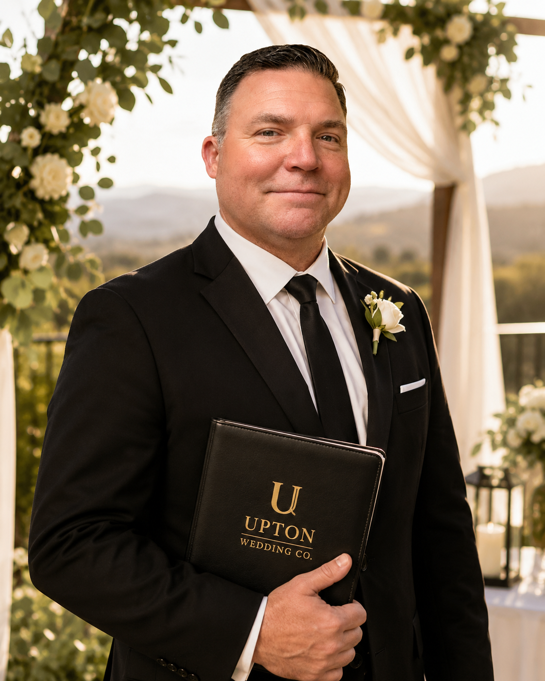 A man in a black suit and tie holding a black folder with gold lettering, standing outdoors during a wedding ceremony.