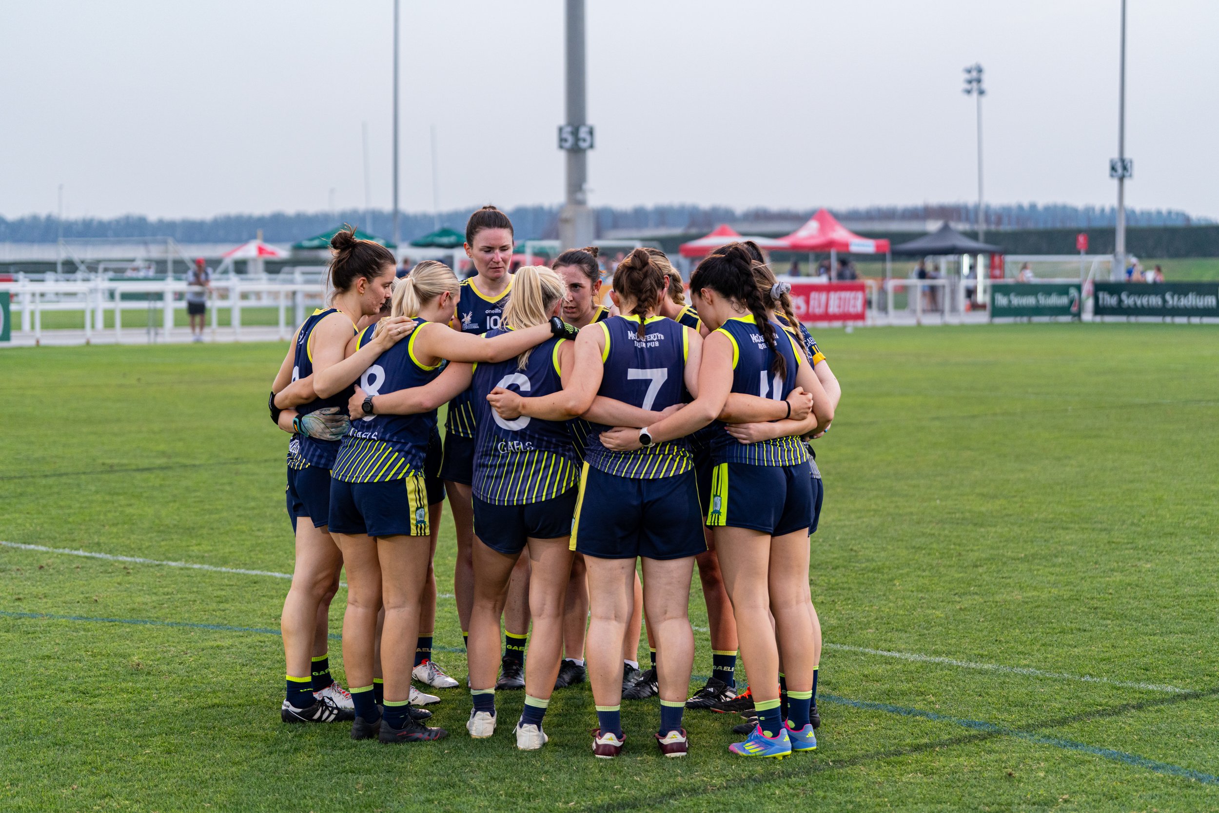 Women’s rugby team huddling on a grassy field during a game, wearing blue and yellow uniforms, with tents and stadium lights in the background.