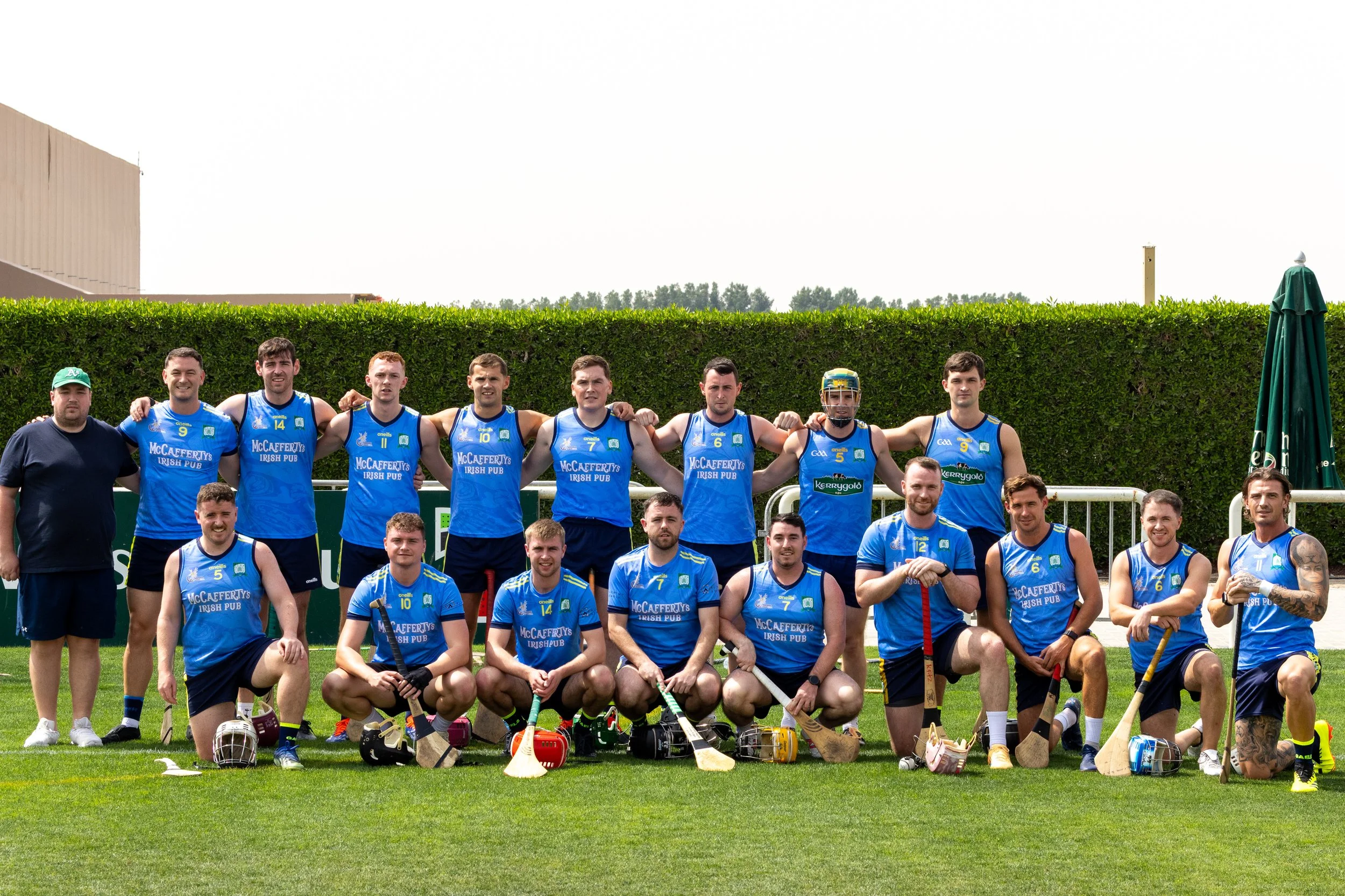 Group of male hurling players and coach standing on a grass field, posing for a team photo. The players are wearing blue uniforms with sponsors' logos, and some are holding field hockey sticks.