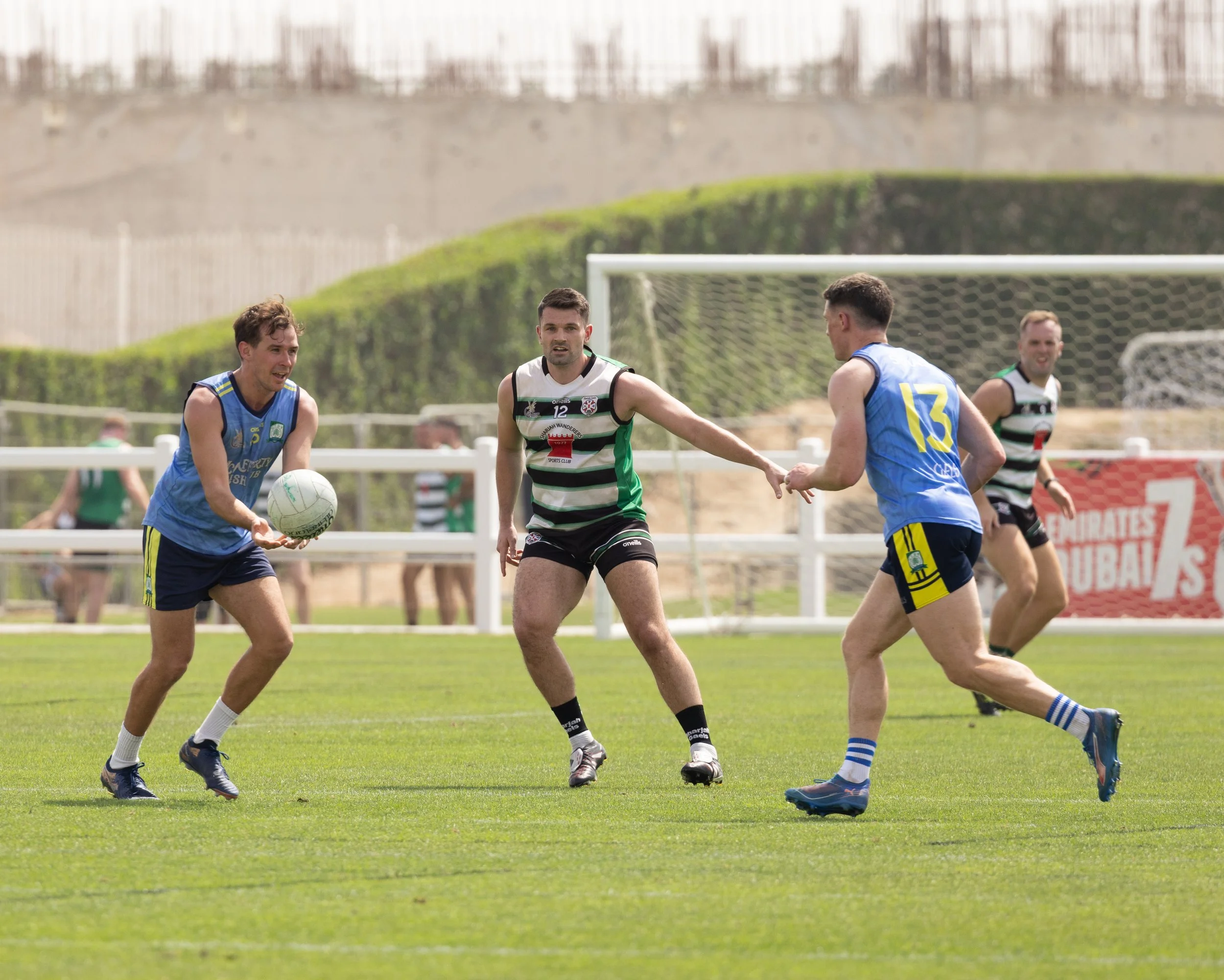 Four male gaelic football players on a green field, one in blue uniform catching the ball while others in black and white striped uniforms attempt to block or defend.