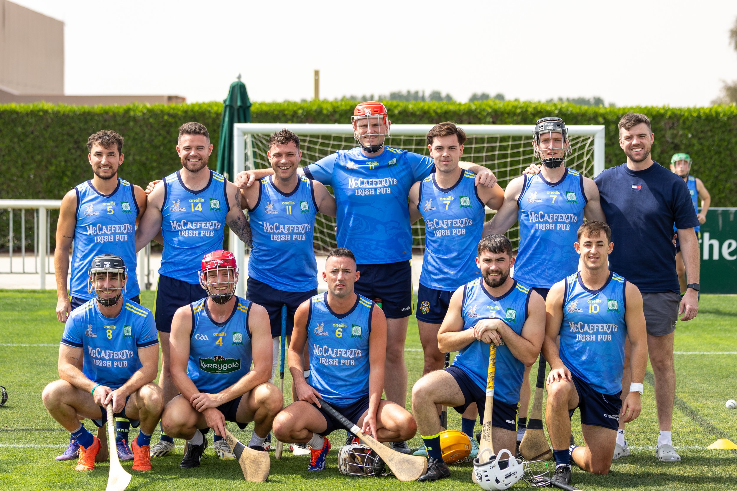 Group of male hurling players and their coach standing on a field in front of a goalpost, wearing blue jerseys with team logos, some with helmets, holding field hockey sticks, smiling for a team photo.