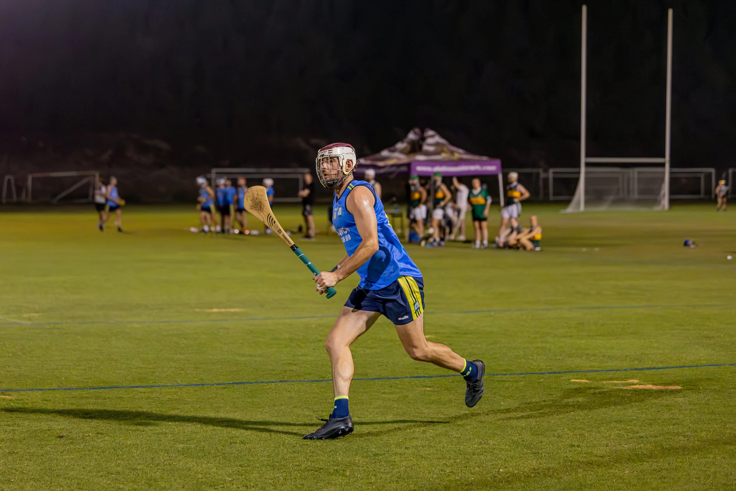 A male field hurling player in a blue uniform and protective helmet running on a grass field at night, with other players and goalposts visible in the background.