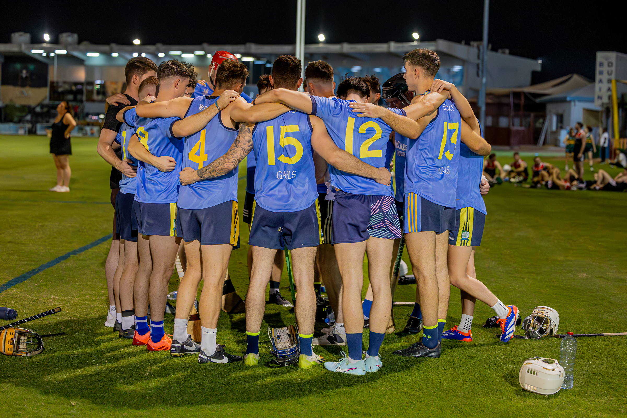 A group of male hurling players wearing blue jerseys and shorts embrace in a huddle on a grassy field night. Helmets and equipment are on the ground nearby.
