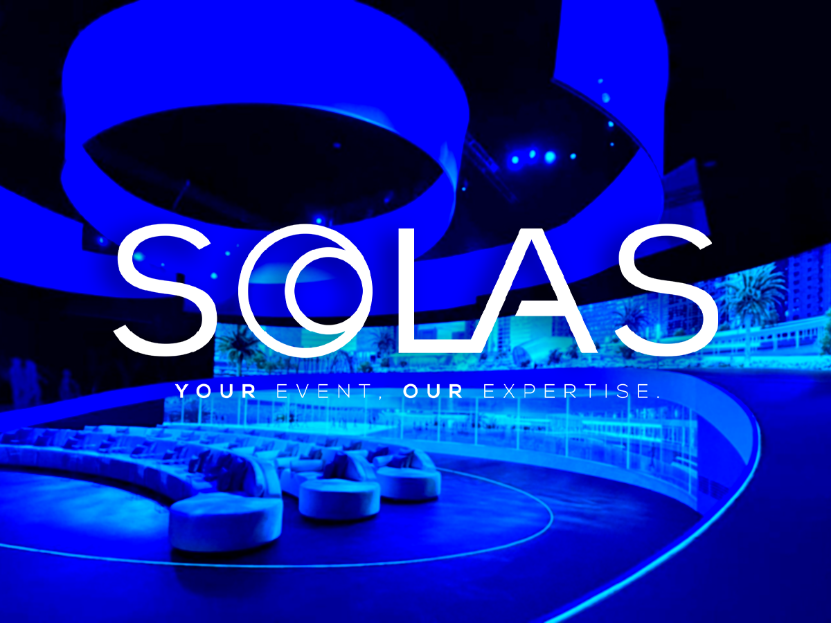 A modern, blue-lit event space with curved seating and a bar area. The features include large hanging circular structures and a backdrop displaying abstract designs. The text overlay reads 'SOLAS' and 'Your Event, Our Expertise'.