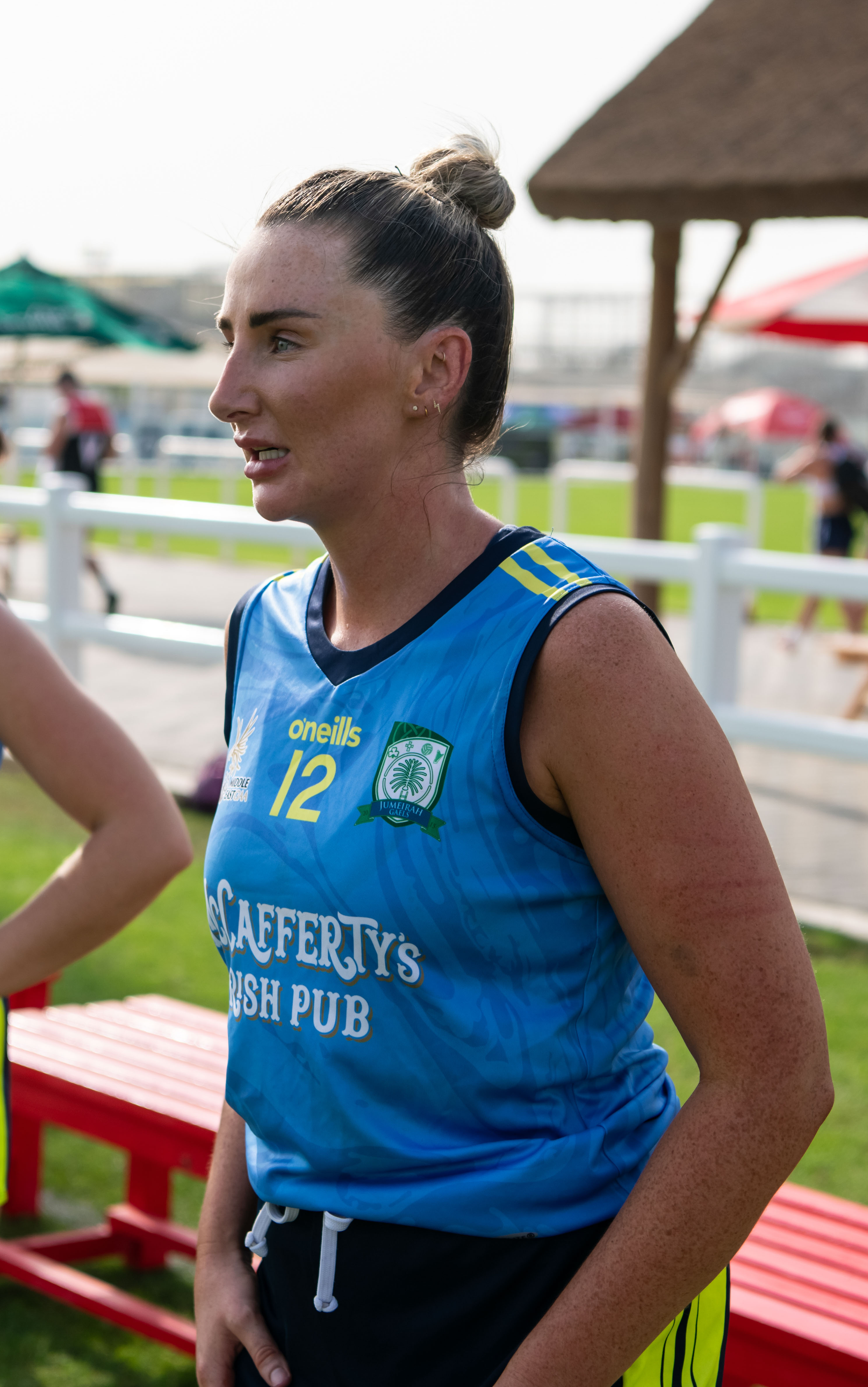 A woman with dark hair tied in a bun wearing a blue sports jersey, standing outdoors near a red bench and under a thatched umbrella. She has a focused expression and is participating in an activity, with a background of blurred people, green grass, and sky.