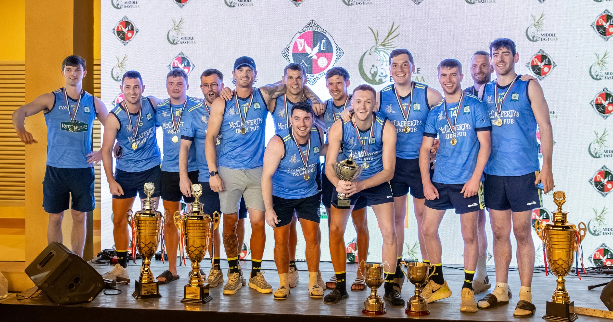 A group of young men in blue sports uniforms celebrating on a stage with trophies, medals, and a trophy in the center, after a sports competition.